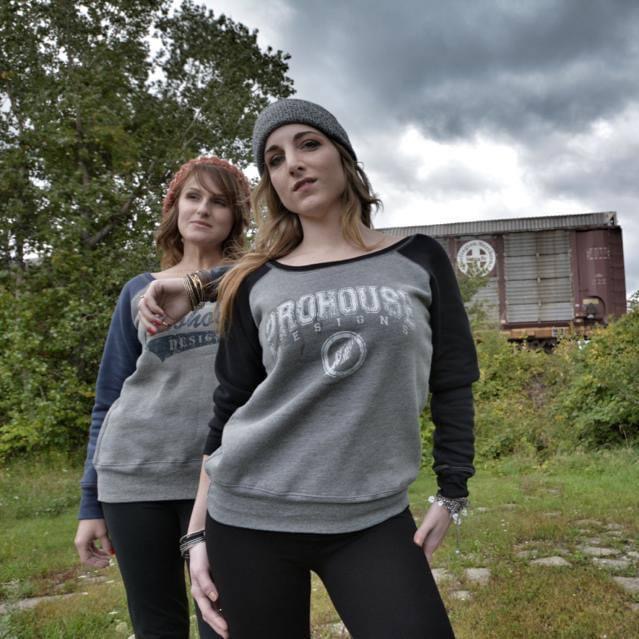 Two women standing outdoors with trees and a train in the background, one wearing a gray beanie and sweatshirt, the other in a gray and black sweatshirt.