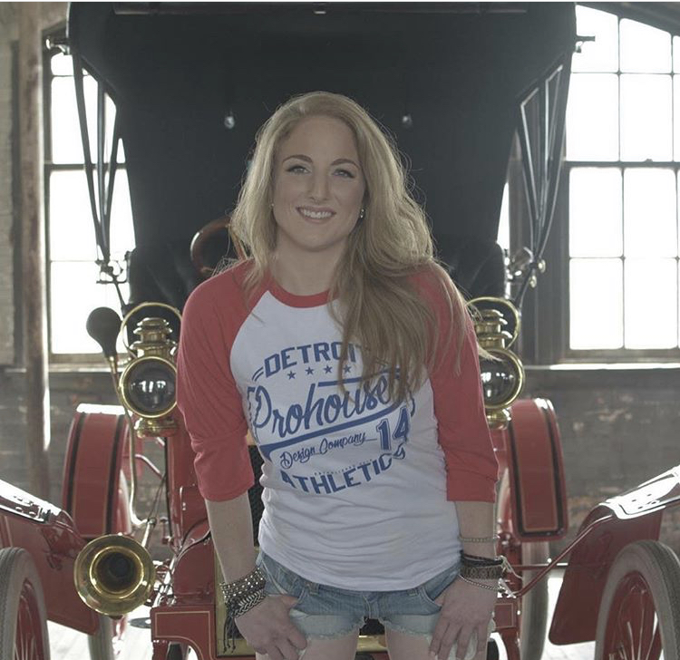 Woman with long blonde hair smiling, wearing a red and white baseball-style shirt, standing in front of a vintage vehicle.