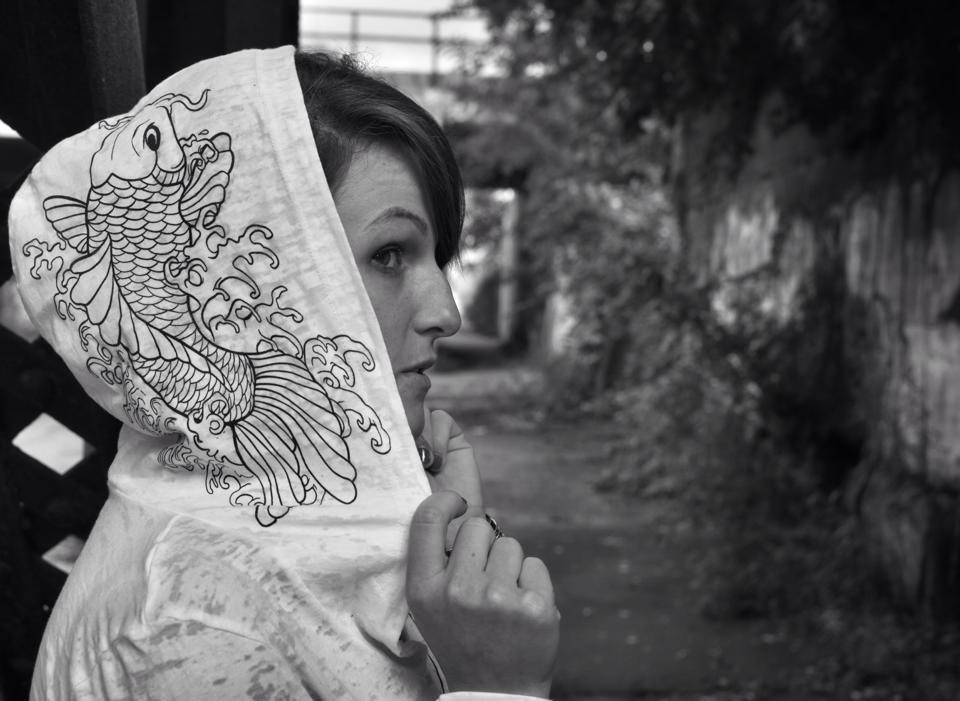 Woman holding a fan with a fish design, standing outdoors with trees and a pathway in the background.
