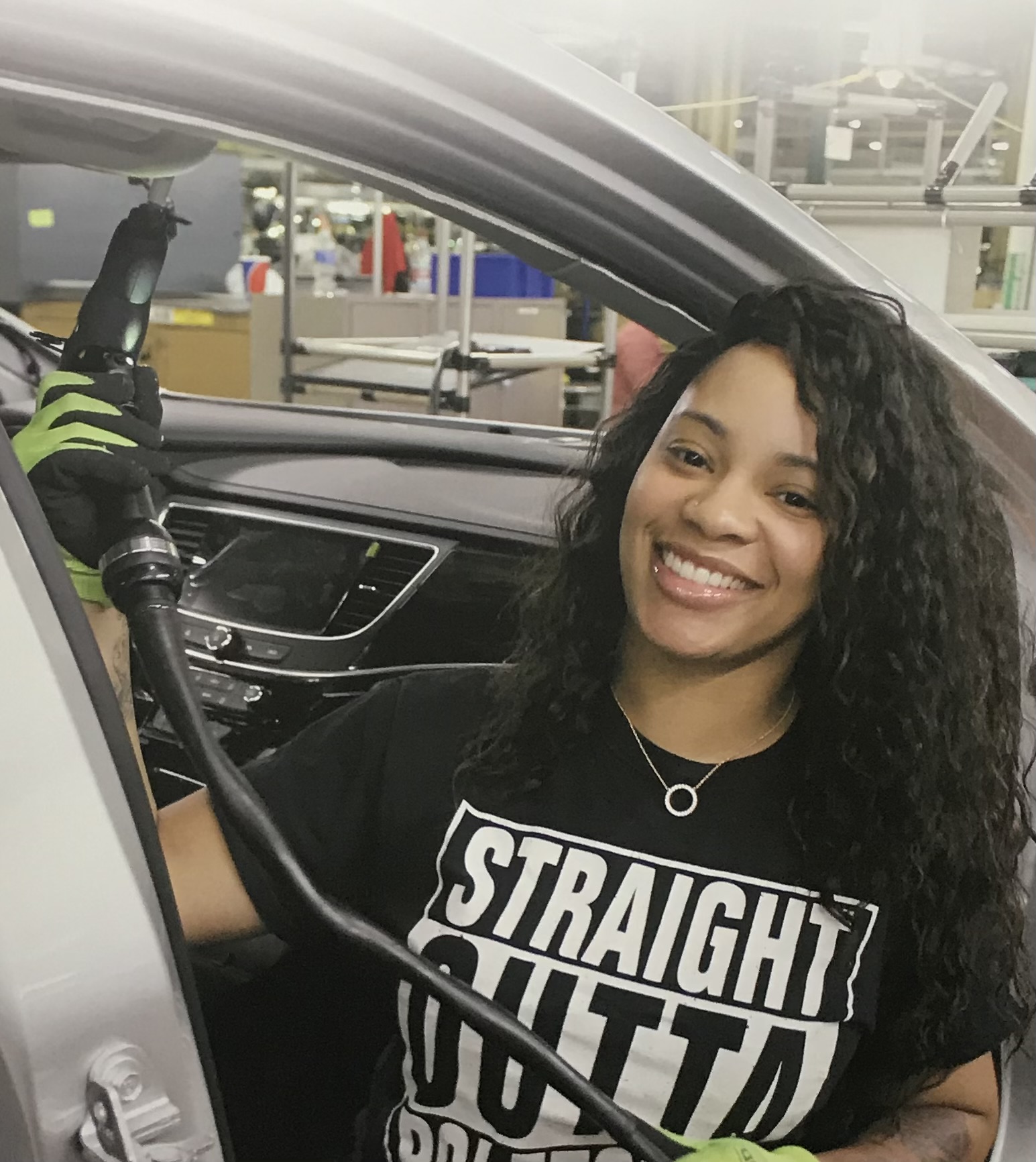Smiling woman with curly hair sitting in a vehicle, wearing a black T-shirt with white text, inside a factory or workshop.