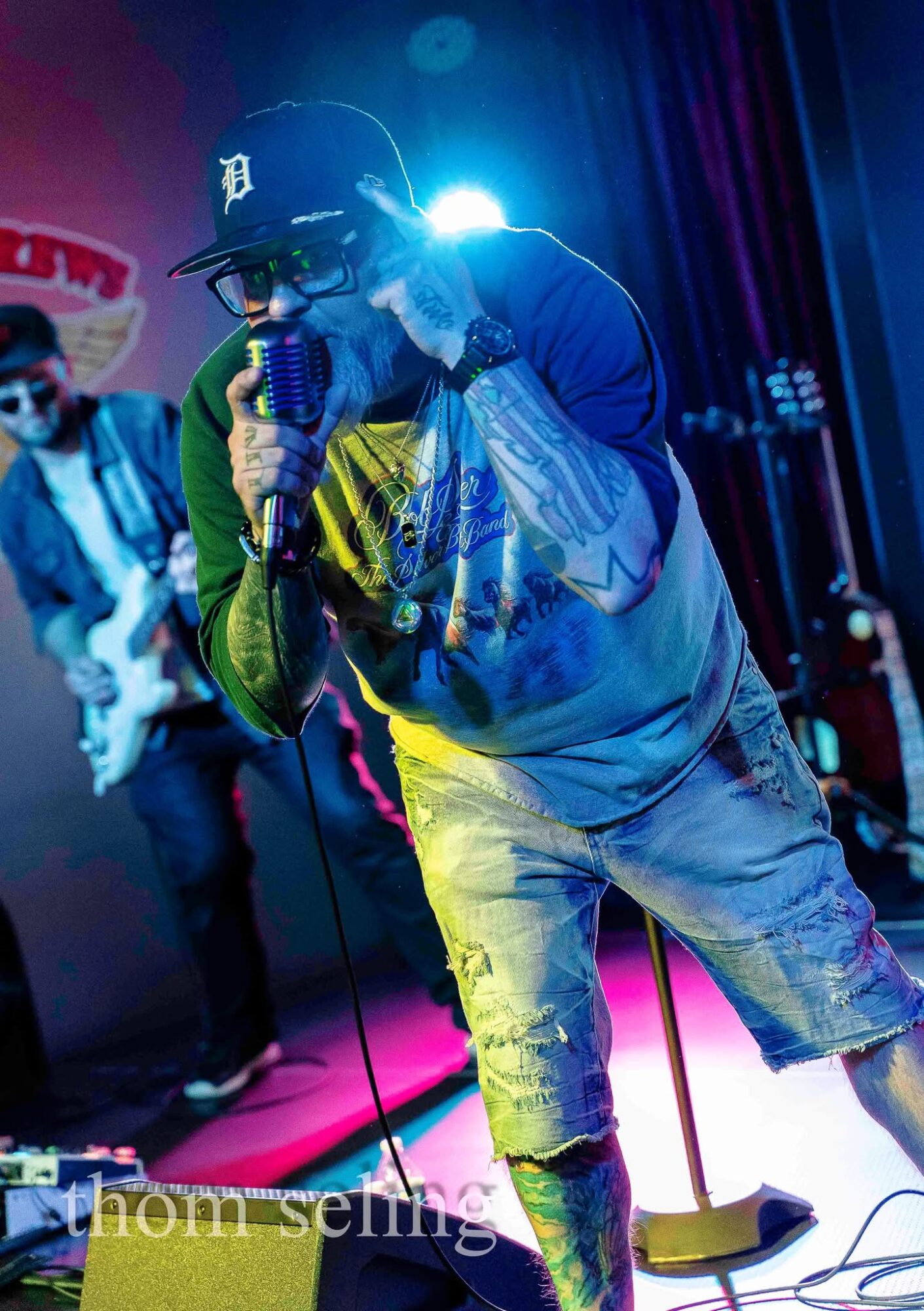 Performer singing into microphone on stage with band, colorful lighting, wearing cap and casual clothing.
