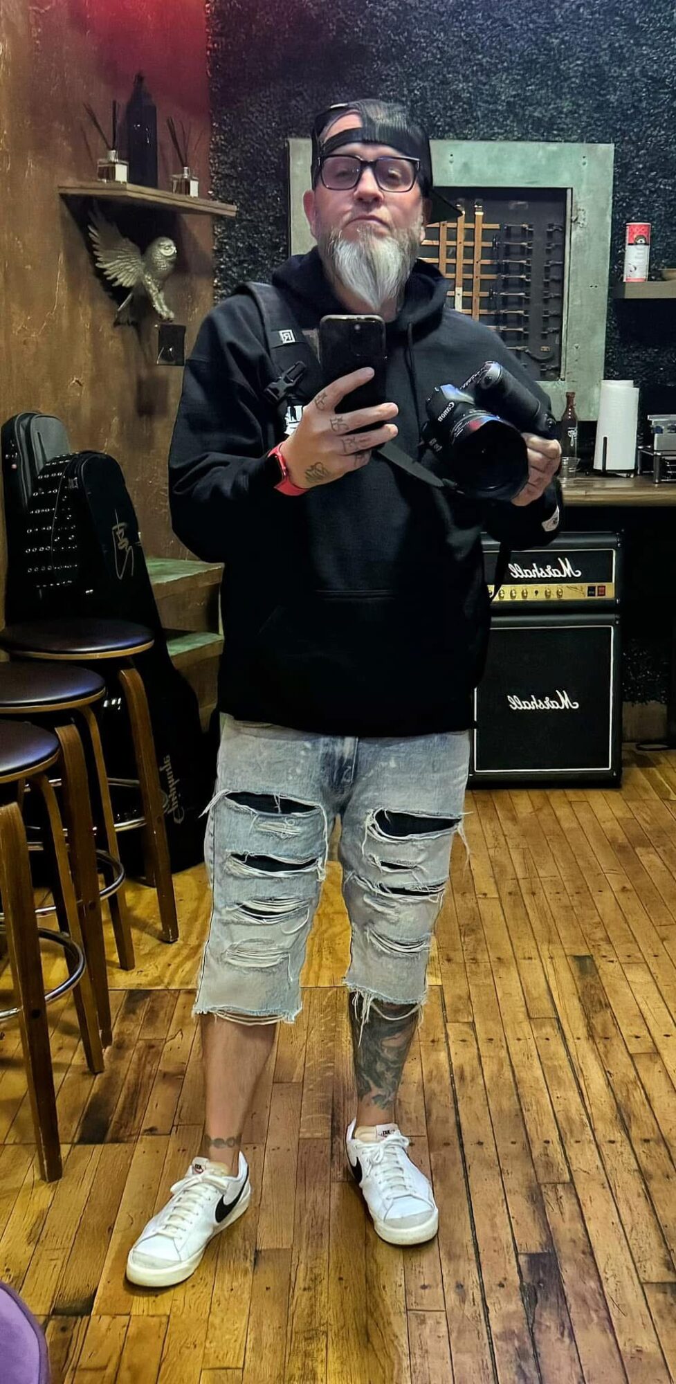 Person taking a mirror selfie in a room with wooden floors, chairs, and musical equipment, wearing a black jacket, shorts, and sneakers.