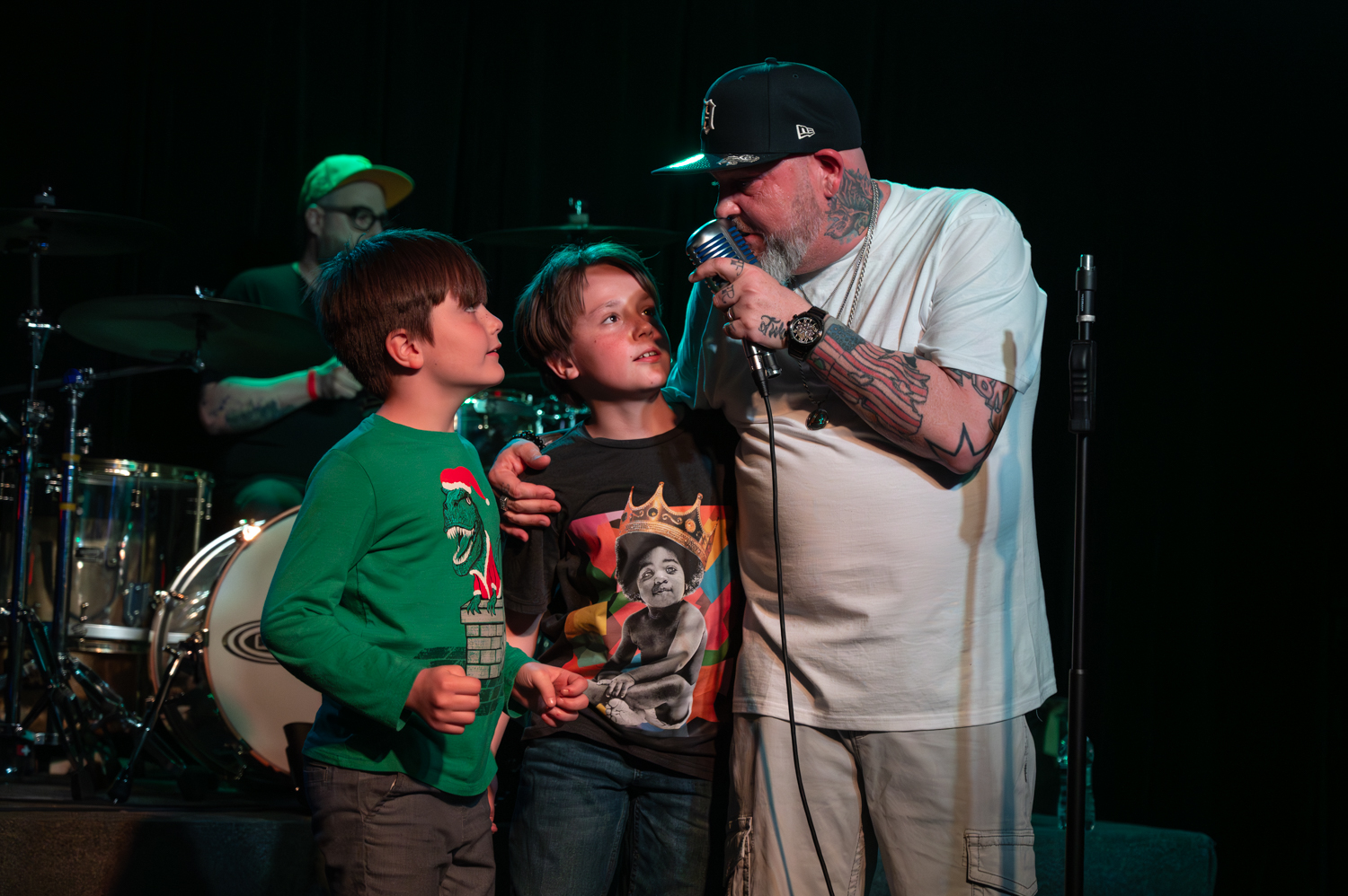 Man with tattoos holding microphone, talking to two children on stage, with a drummer in the background.