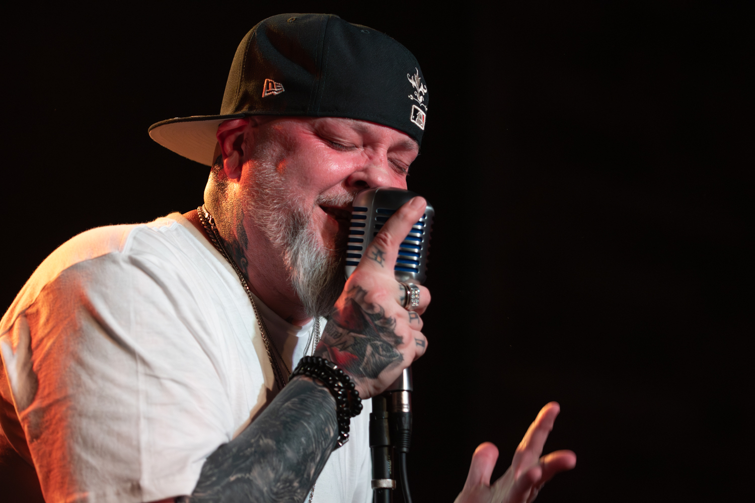 Man with a beard and tattoos singing into a microphone, wearing a black cap and white shirt, against a dark background.