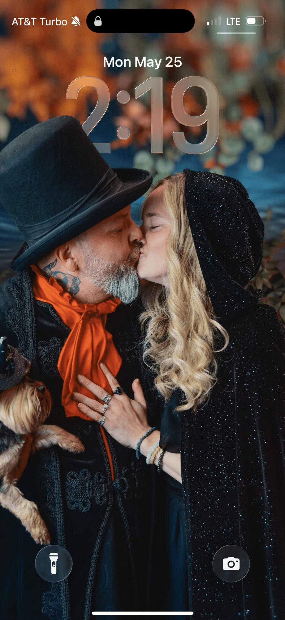 Two people, a man and a woman, face each other close, outdoors with blurred colorful background, wearing dark clothing and hats.