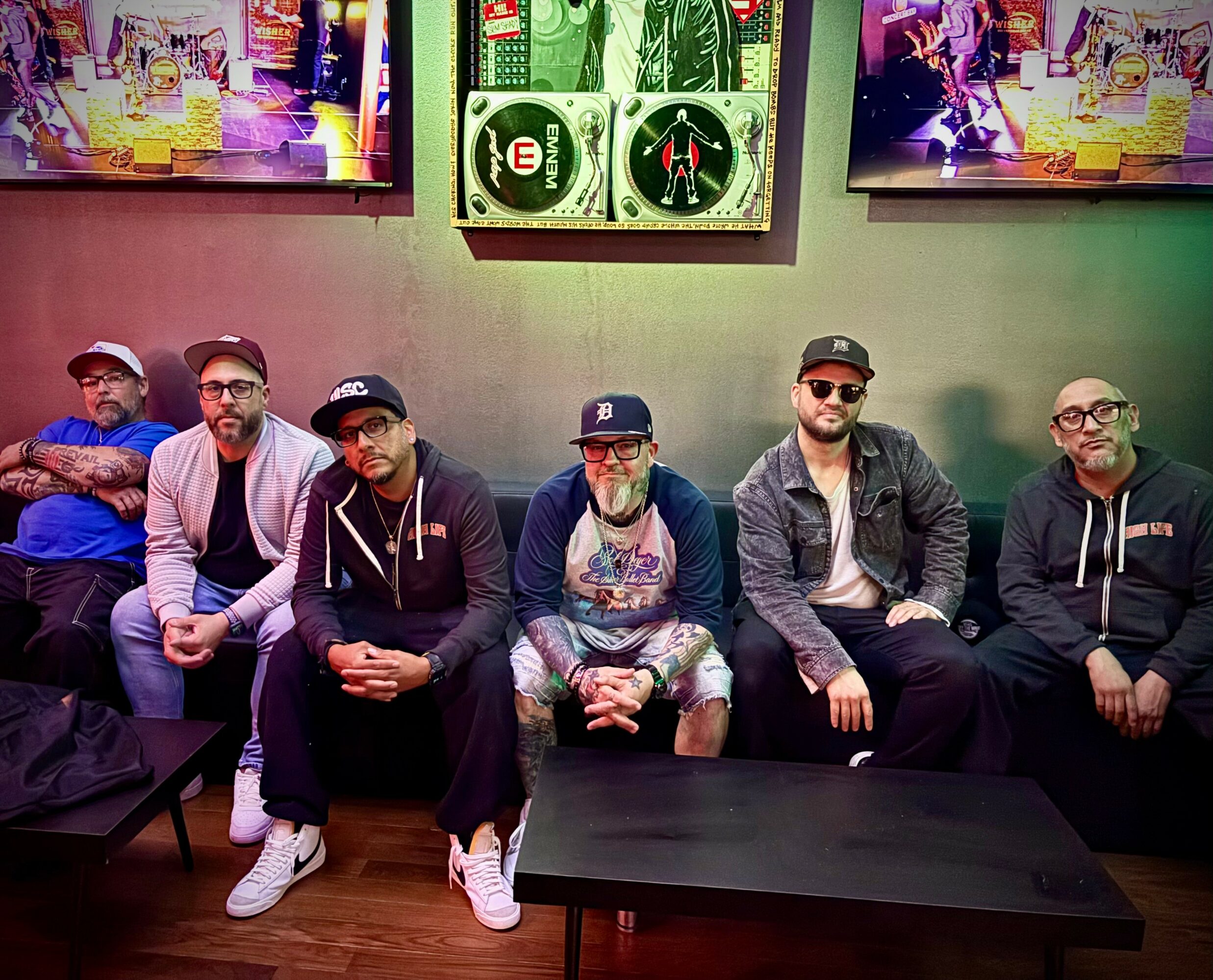 Six men sitting on a bench in a room with colorful wall art and a dartboard, some wearing glasses and hats.