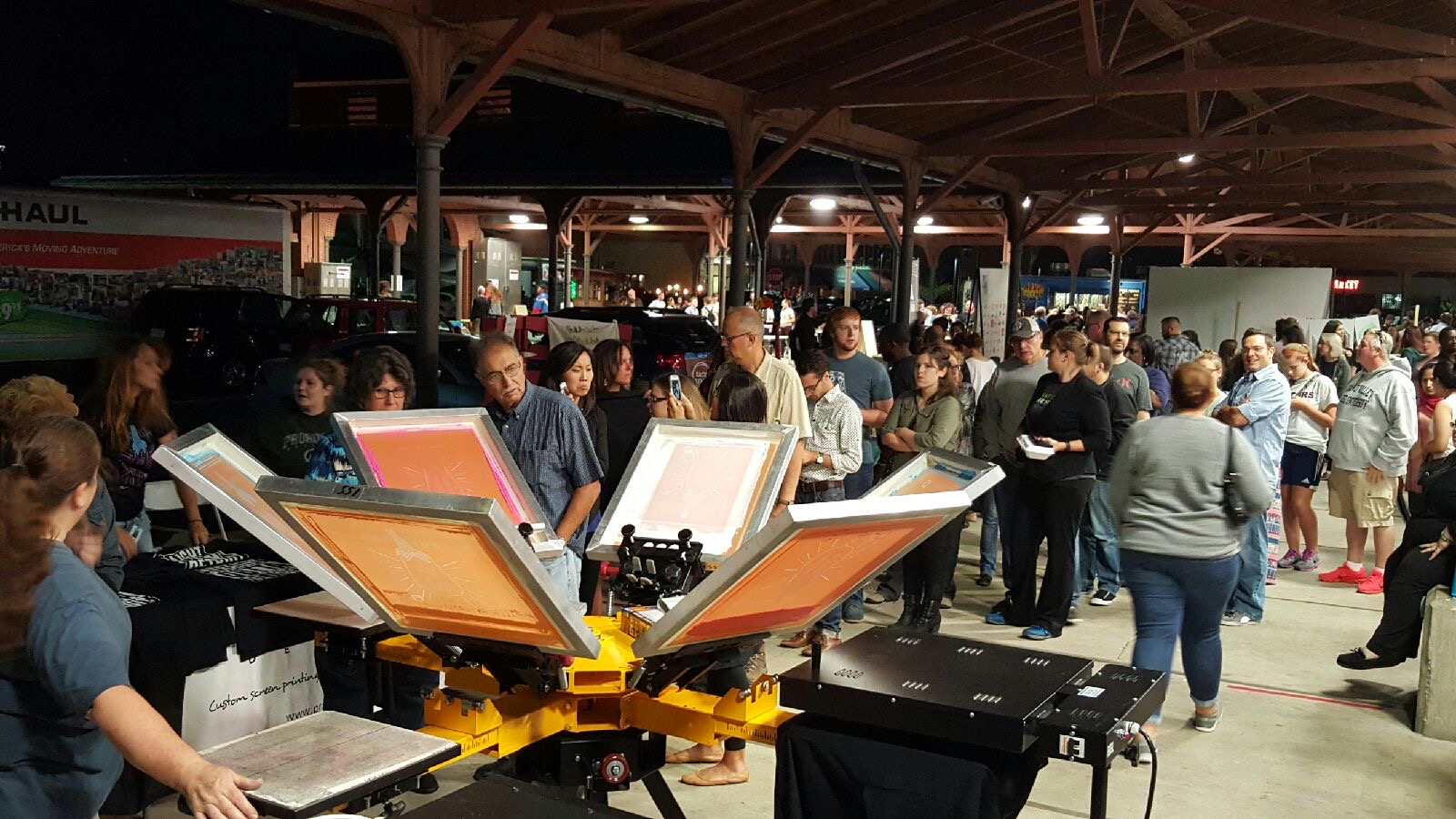 People gather under a large covered area with tables and equipment, some standing and some sitting, in a busy indoor setting.