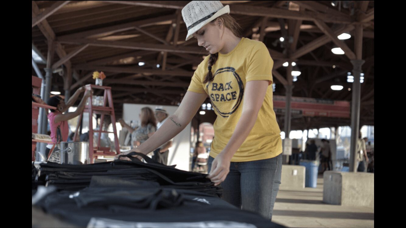 Young person wearing a yellow T-shirt and white cap, browsing items at an outdoor market or fair, with wooden structures in background.