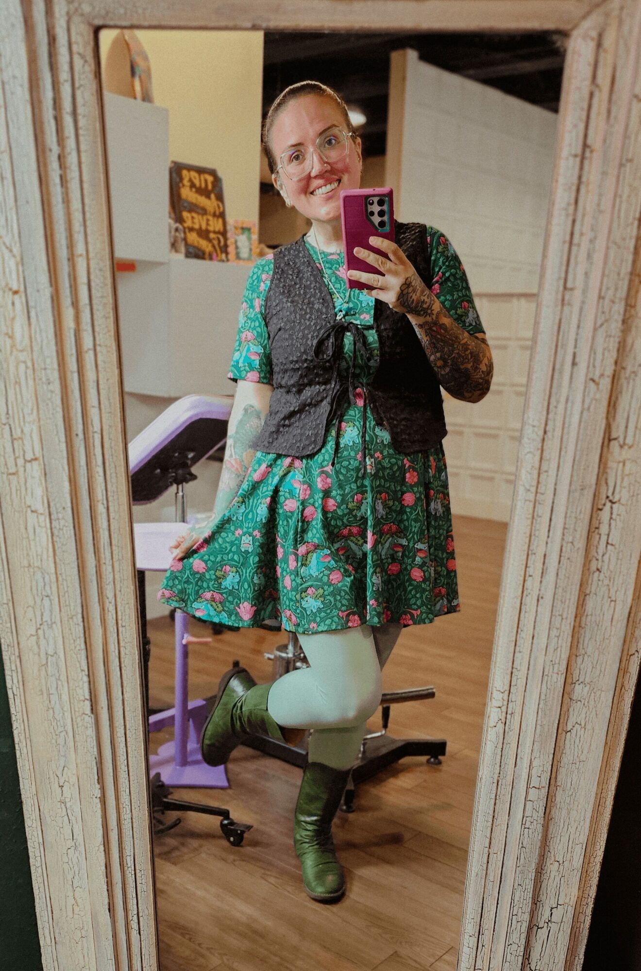 Person taking a mirror selfie, wearing a green floral dress, gray vest, and green boots, smiling with glasses.