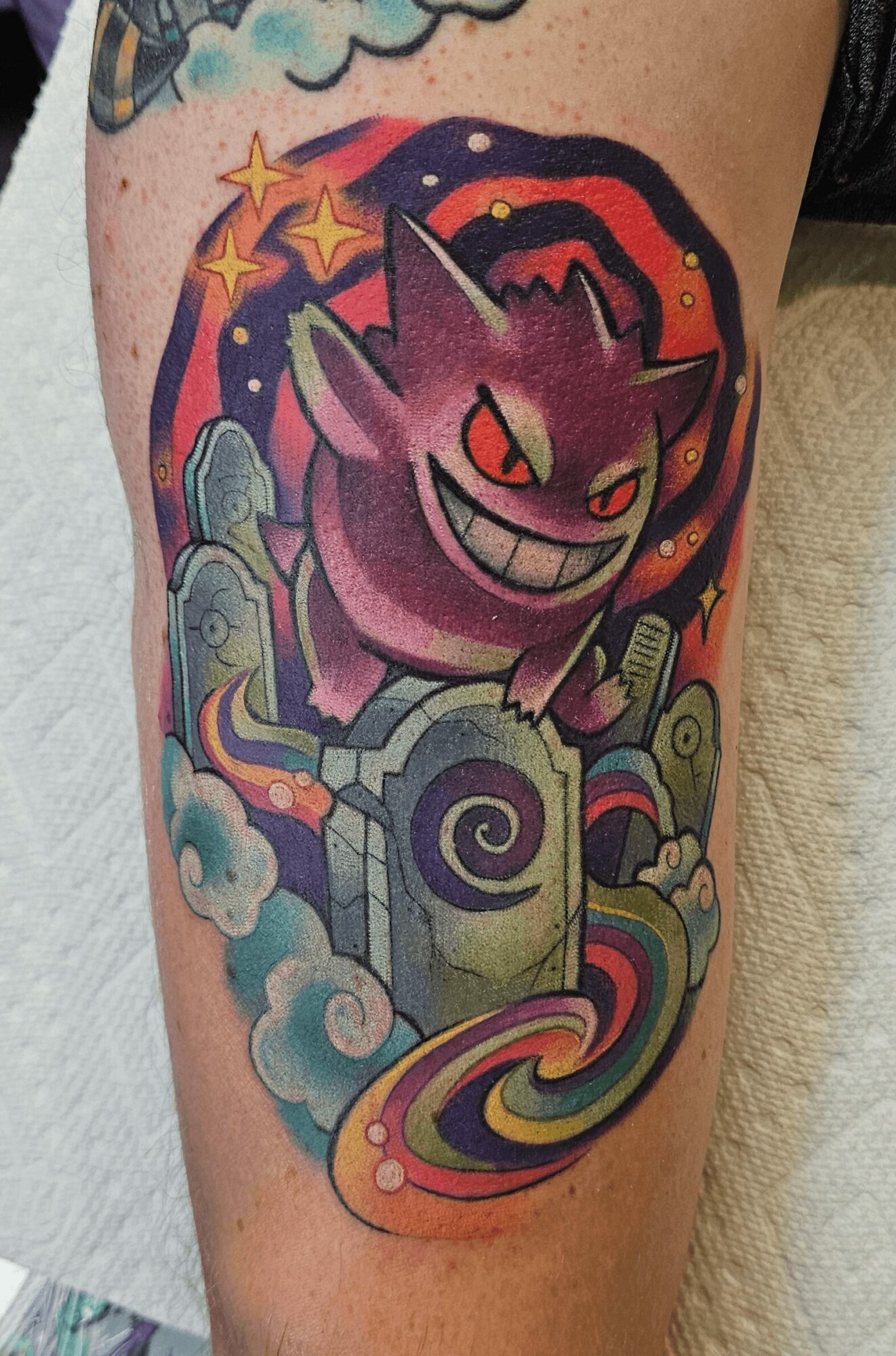 Tattoo of a mischievous purple creature with red eyes, surrounded by colorful clouds, stars, and a swirling rainbow, on a person's arm.