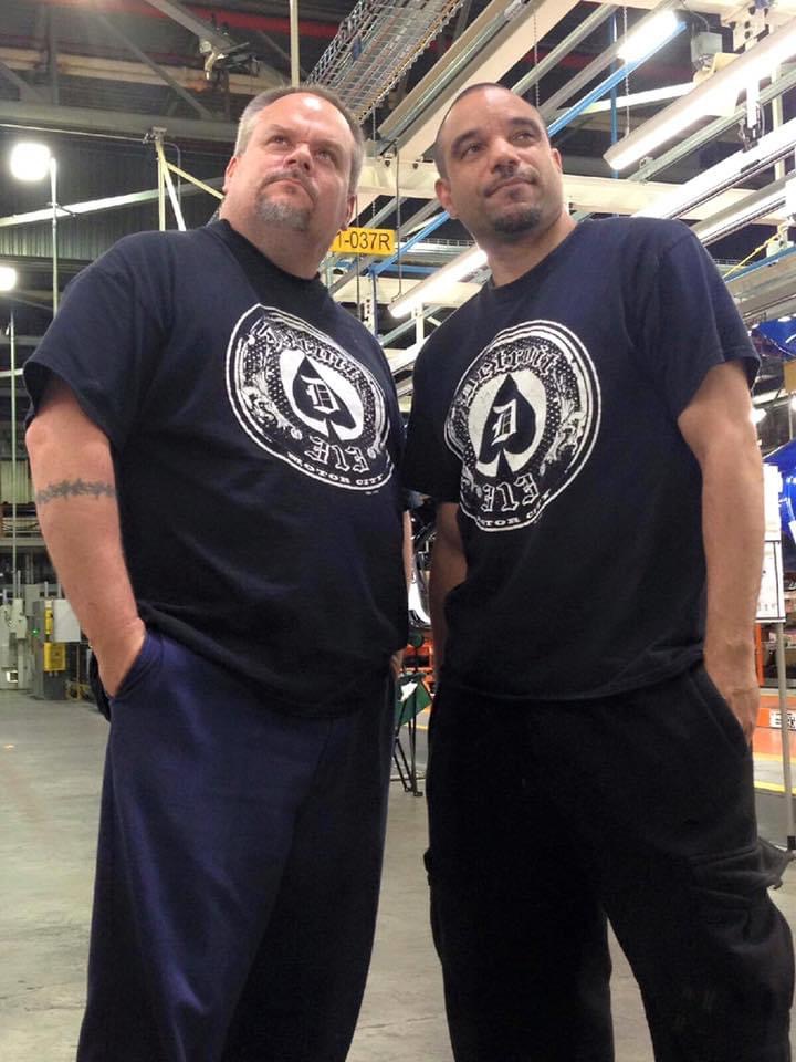 Two men stand side by side in an industrial setting, wearing matching black T-shirts with a circular logo, looking slightly upward.