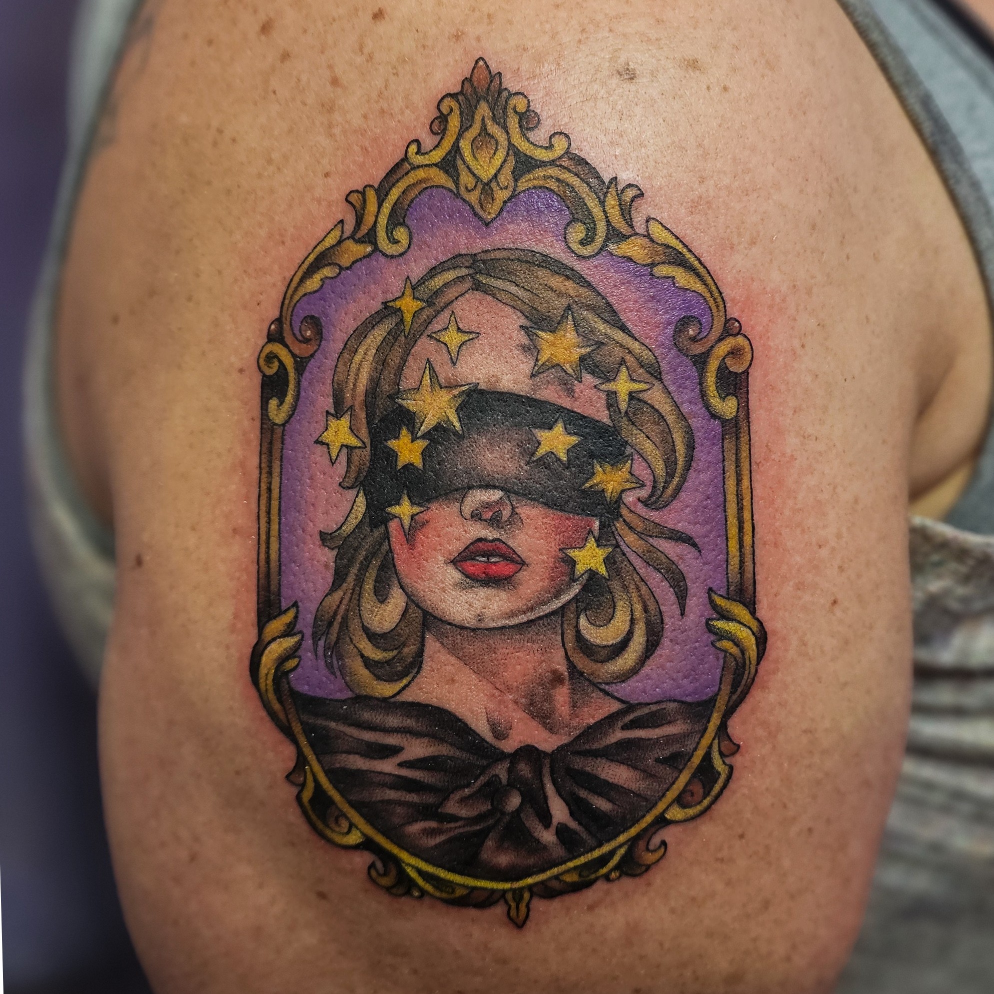 Tattoo of a woman with stars covering her eyes, framed in an ornate mirror with a purple background.