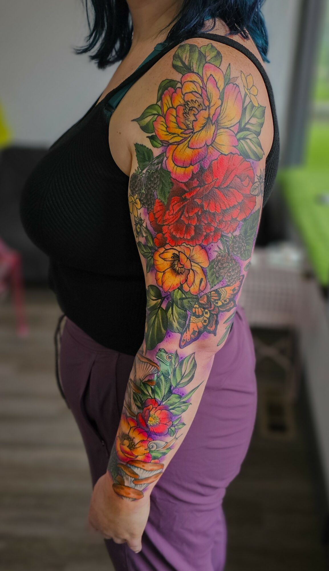 Person with colorful floral tattoo sleeve on arm, wearing black top and purple pants, standing indoors.