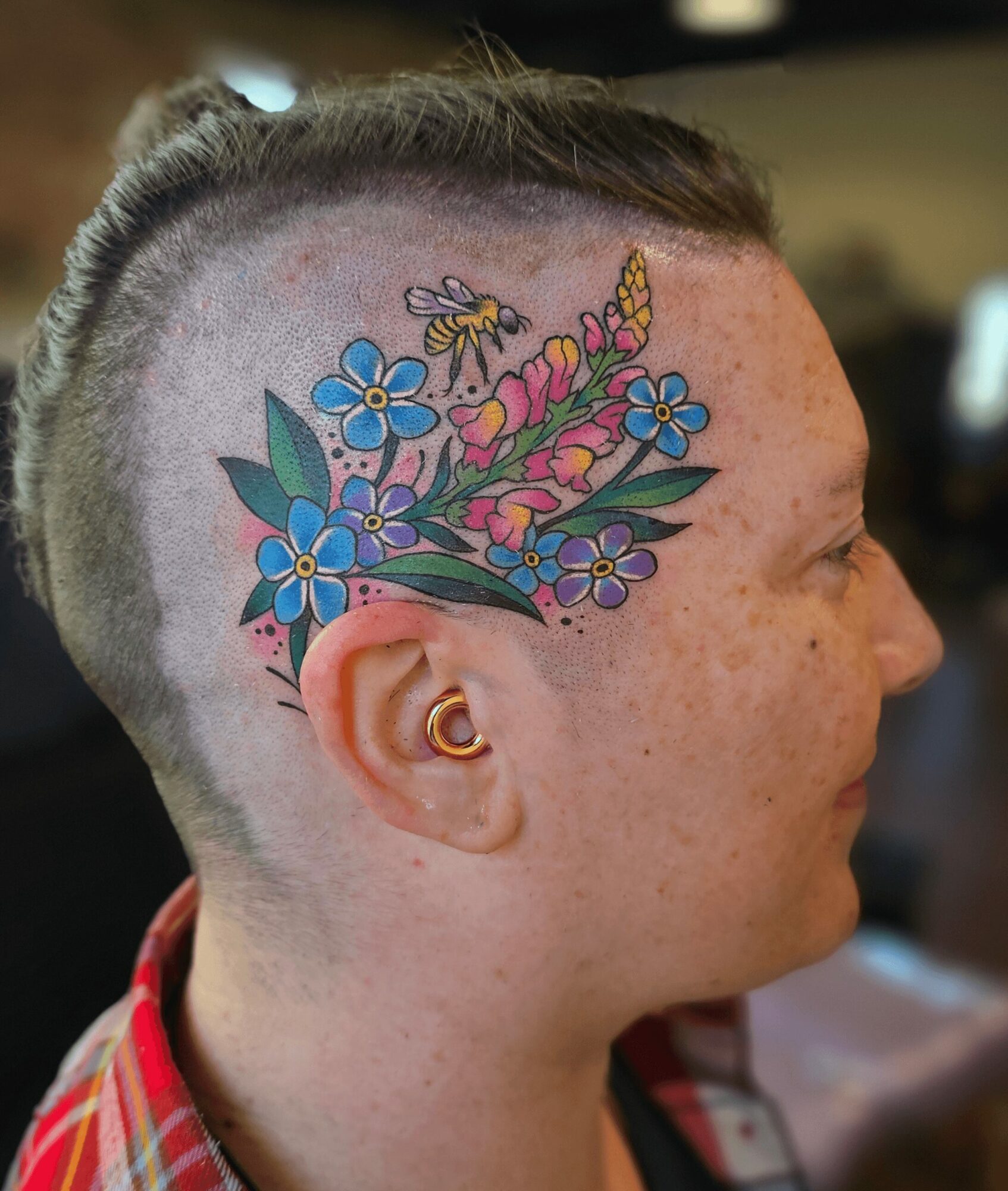 Side view of a person with a colorful floral tattoo on the head, short hair, and a gold ear cuff.