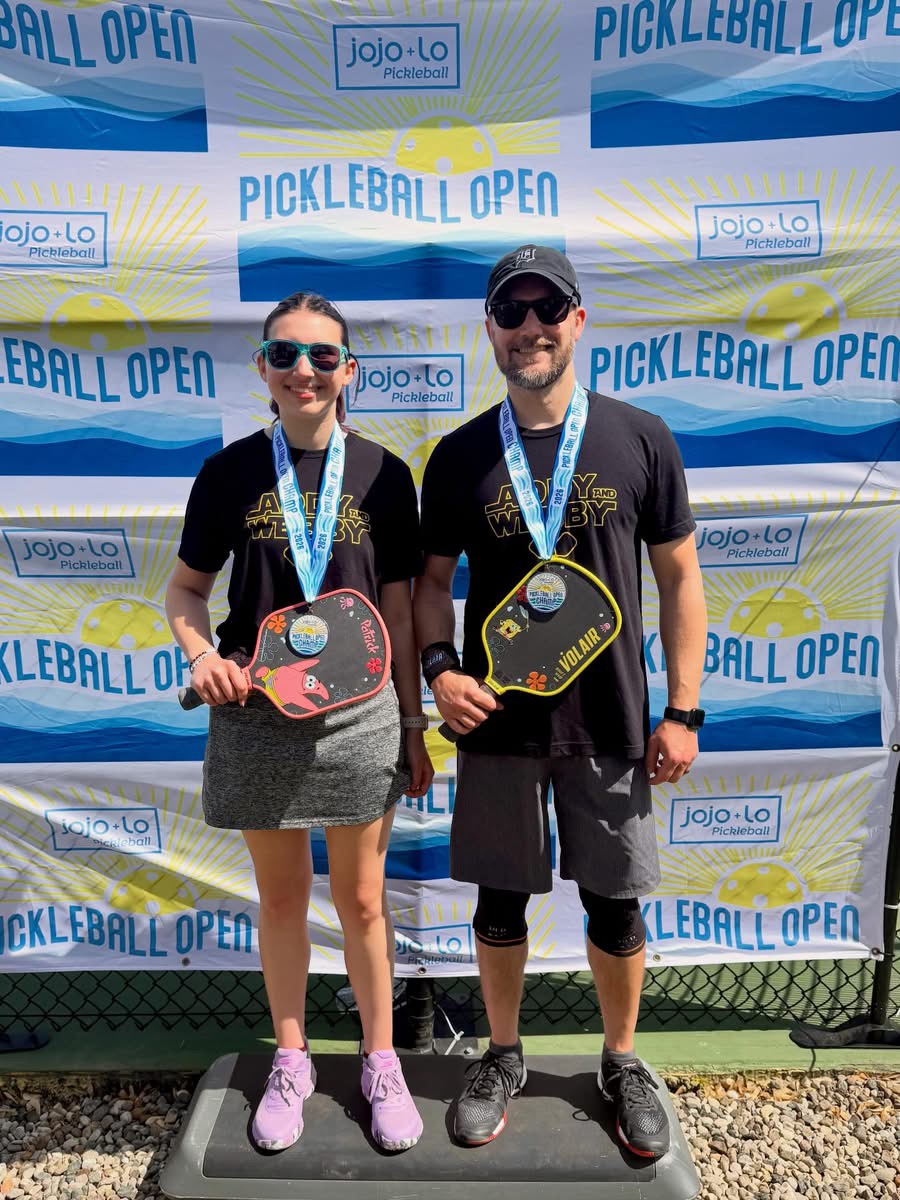 Two people holding pickleball paddles and medals, standing in front of a pickleball open backdrop.