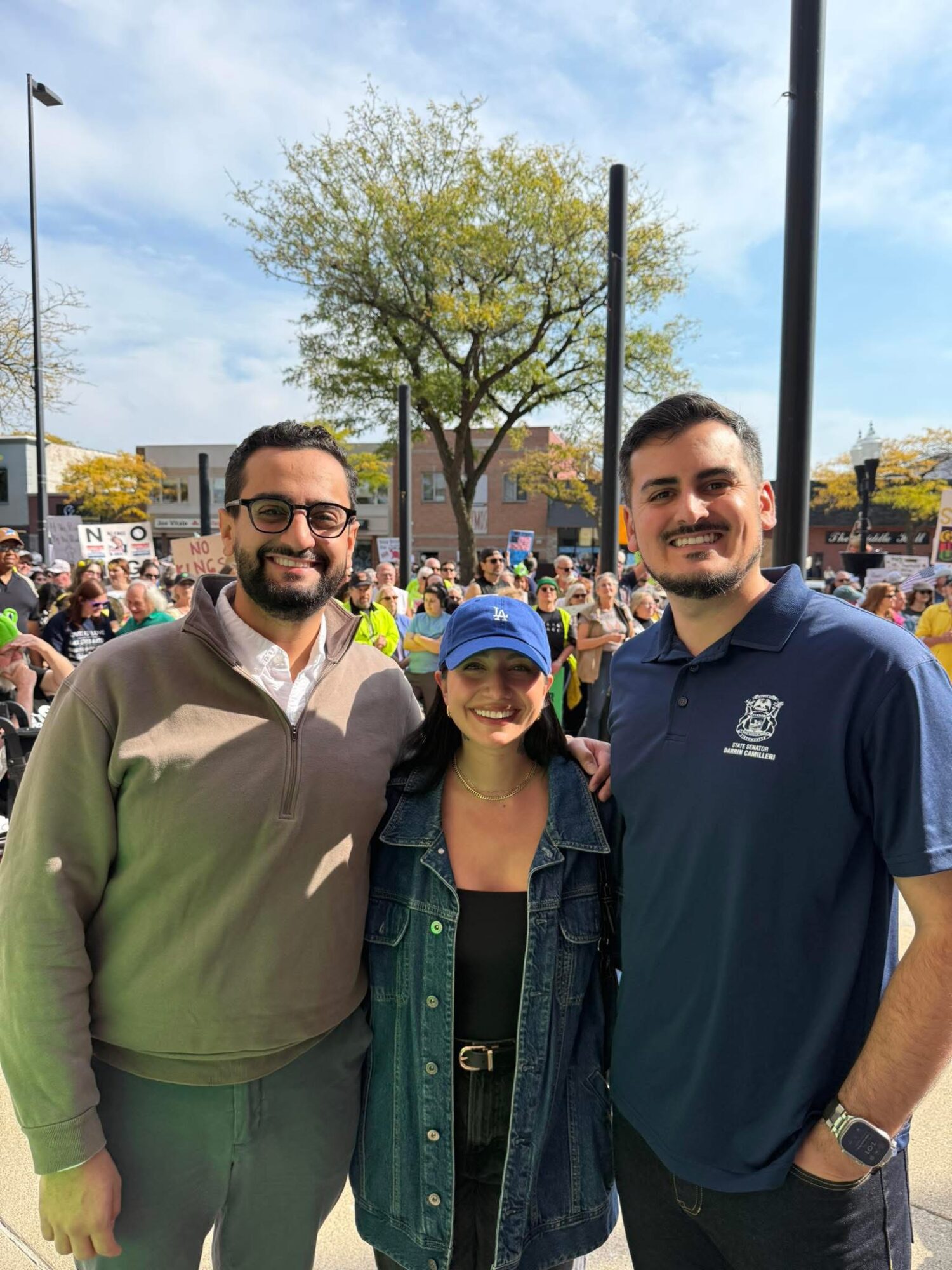 Three people standing outdoors in front of a crowd and trees, smiling at the camera.