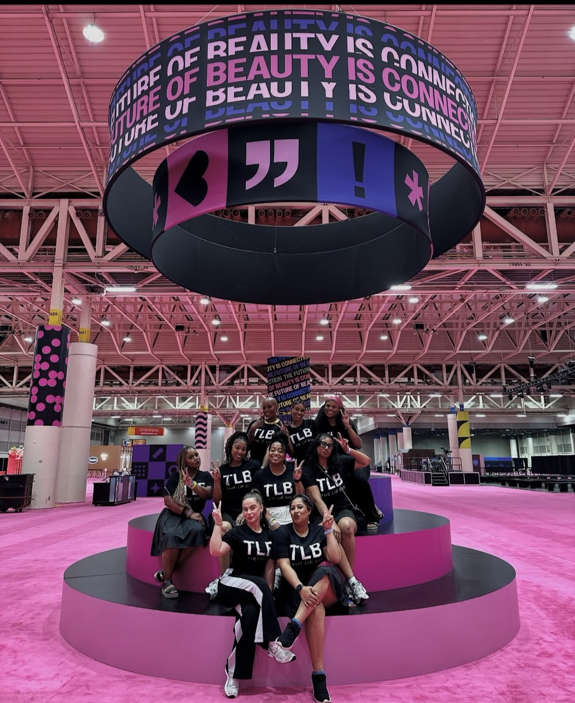 Group of people posing on a pink circular platform inside a large, pink-themed indoor space with a digital display overhead.