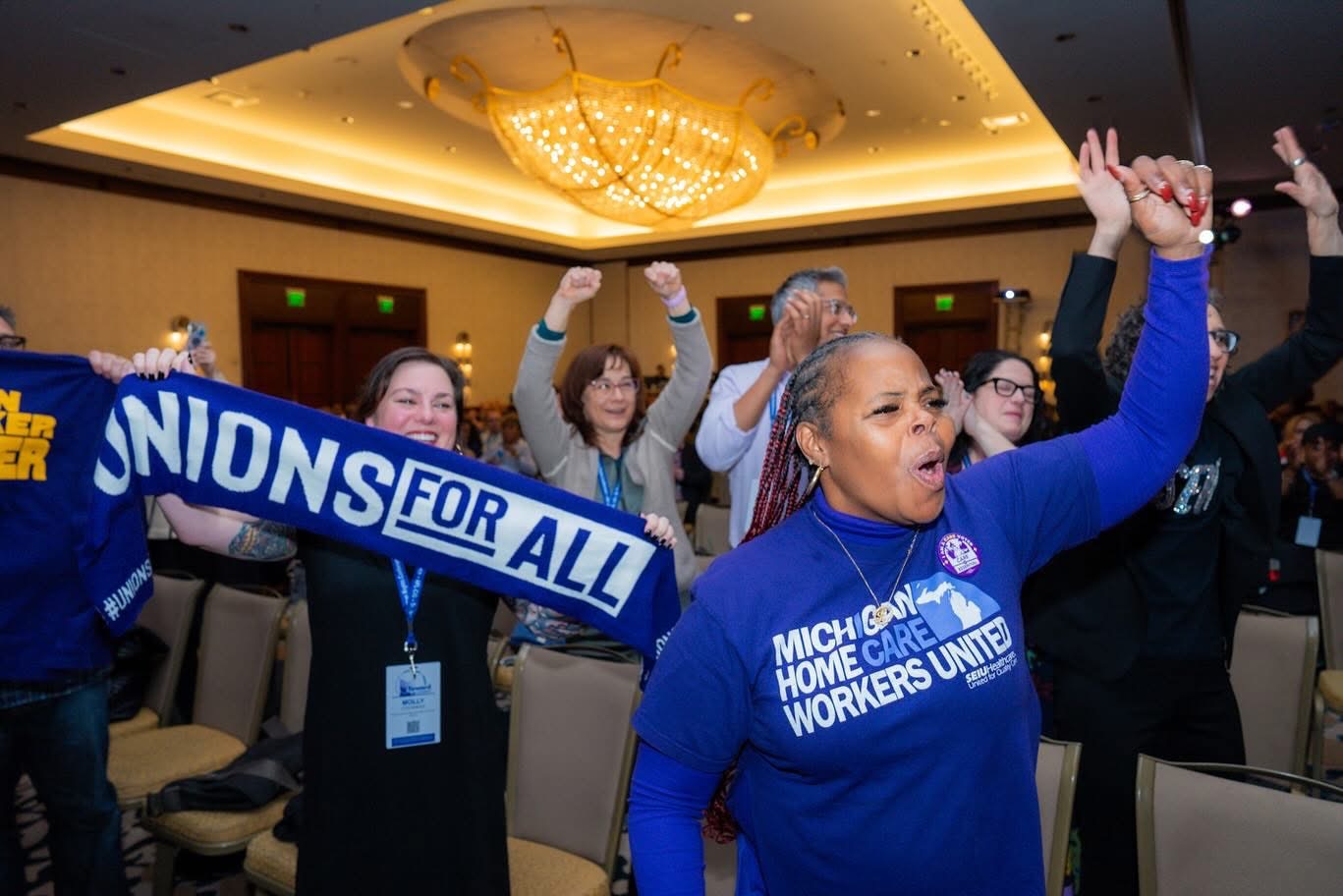 Group of people at a conference or rally, some raising fists, wearing blue shirts, in a large room with a chandelier.