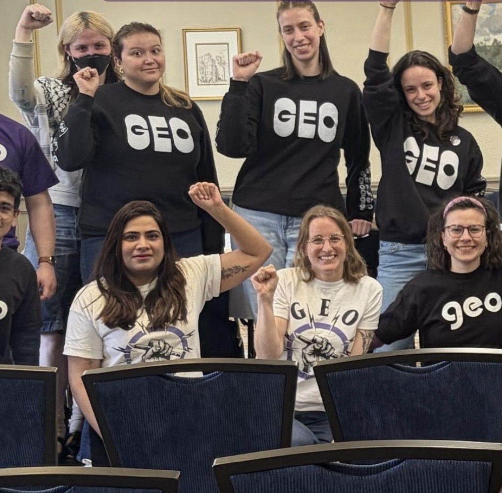 Group of women and girls in a room, some raising fists, wearing black and white GEO shirts, smiling.