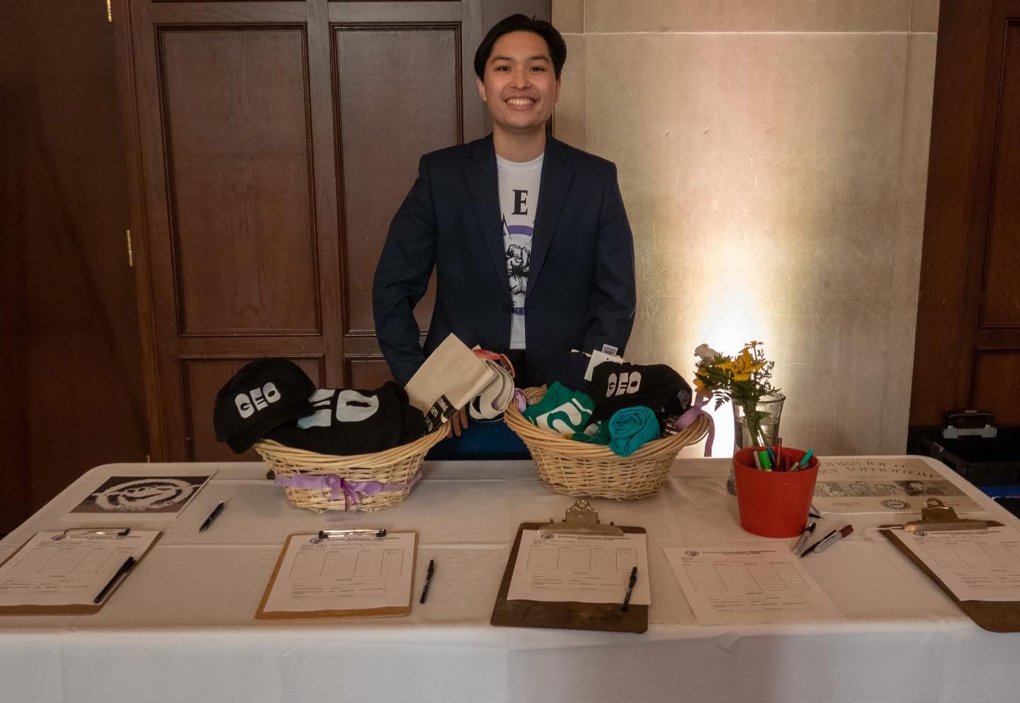 Young man standing behind a table with baskets of socks and a potted plant, smiling, in a wood-paneled room.