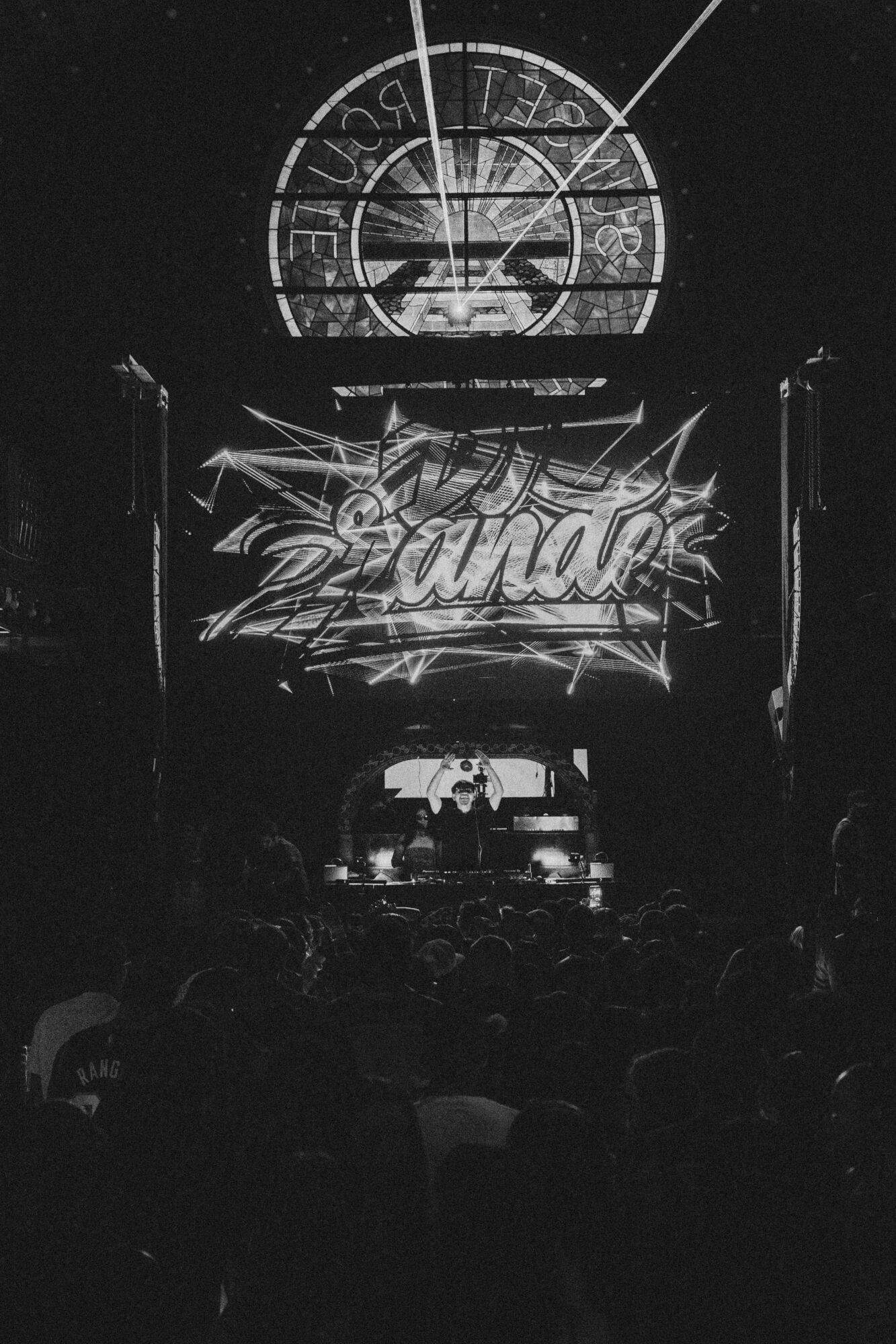 Concert stage with illuminated sign reading 'Canada' and a clock above, audience in foreground, black and white image.