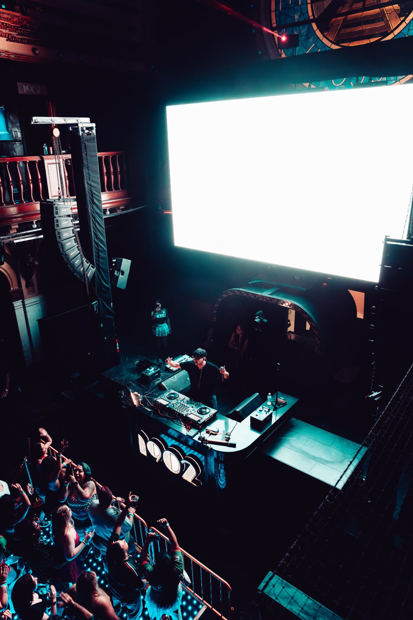 DJ performing on stage with large screen behind, audience watching, dark lighting, electronic equipment visible.