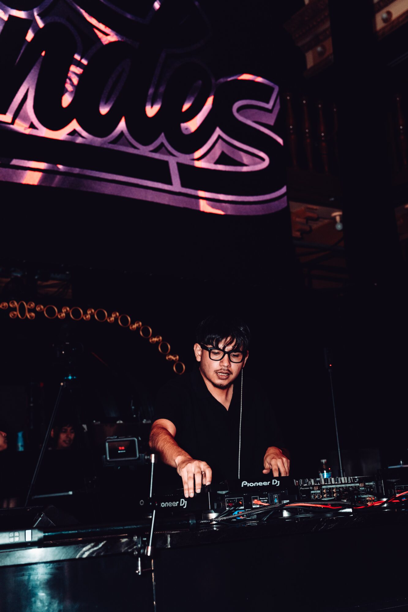 DJ wearing glasses and black shirt performs at a dark nightclub with a large illuminated sign in the background.