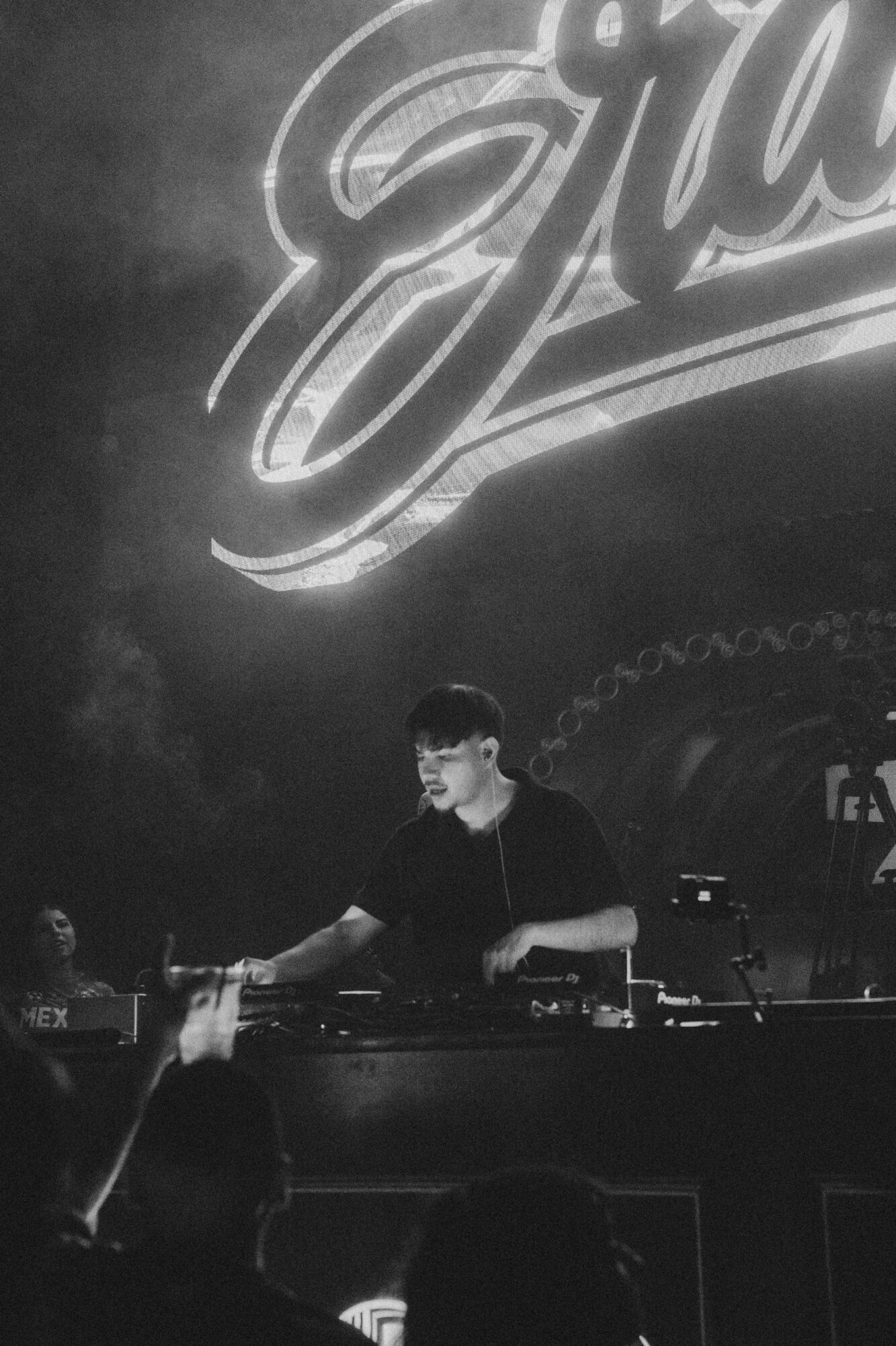 DJ performing on stage with a large illuminated sign in the background, audience in foreground