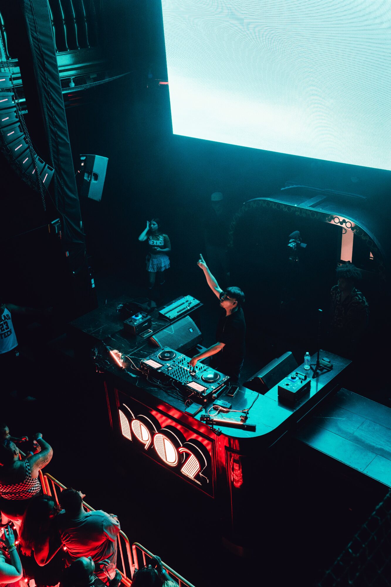 DJ performing at a nightclub with a large screen behind, colorful lighting, and a crowd in front.