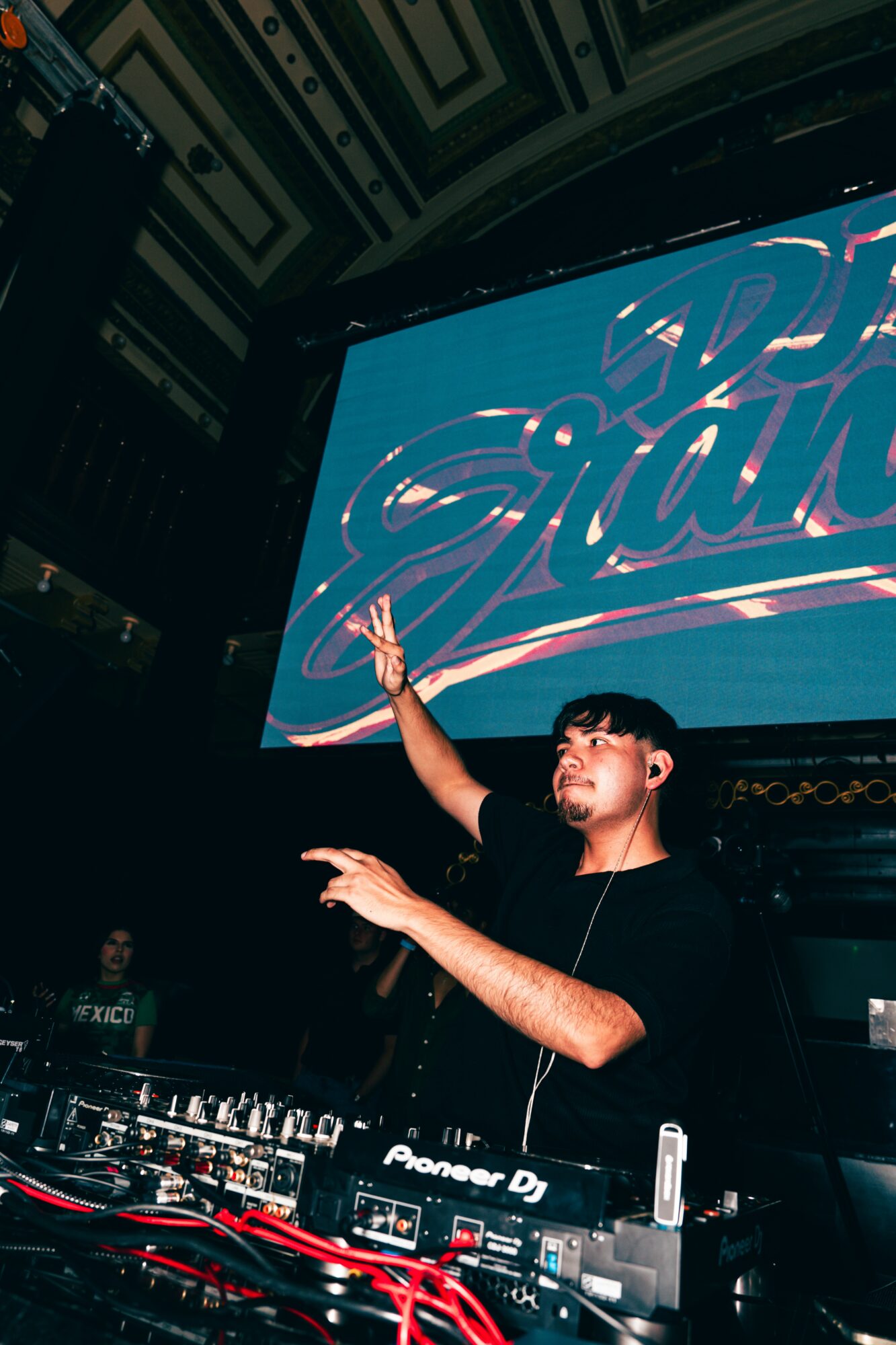 DJ performing on stage with large screen behind displaying stylized text, in a dark venue with equipment and audience.
