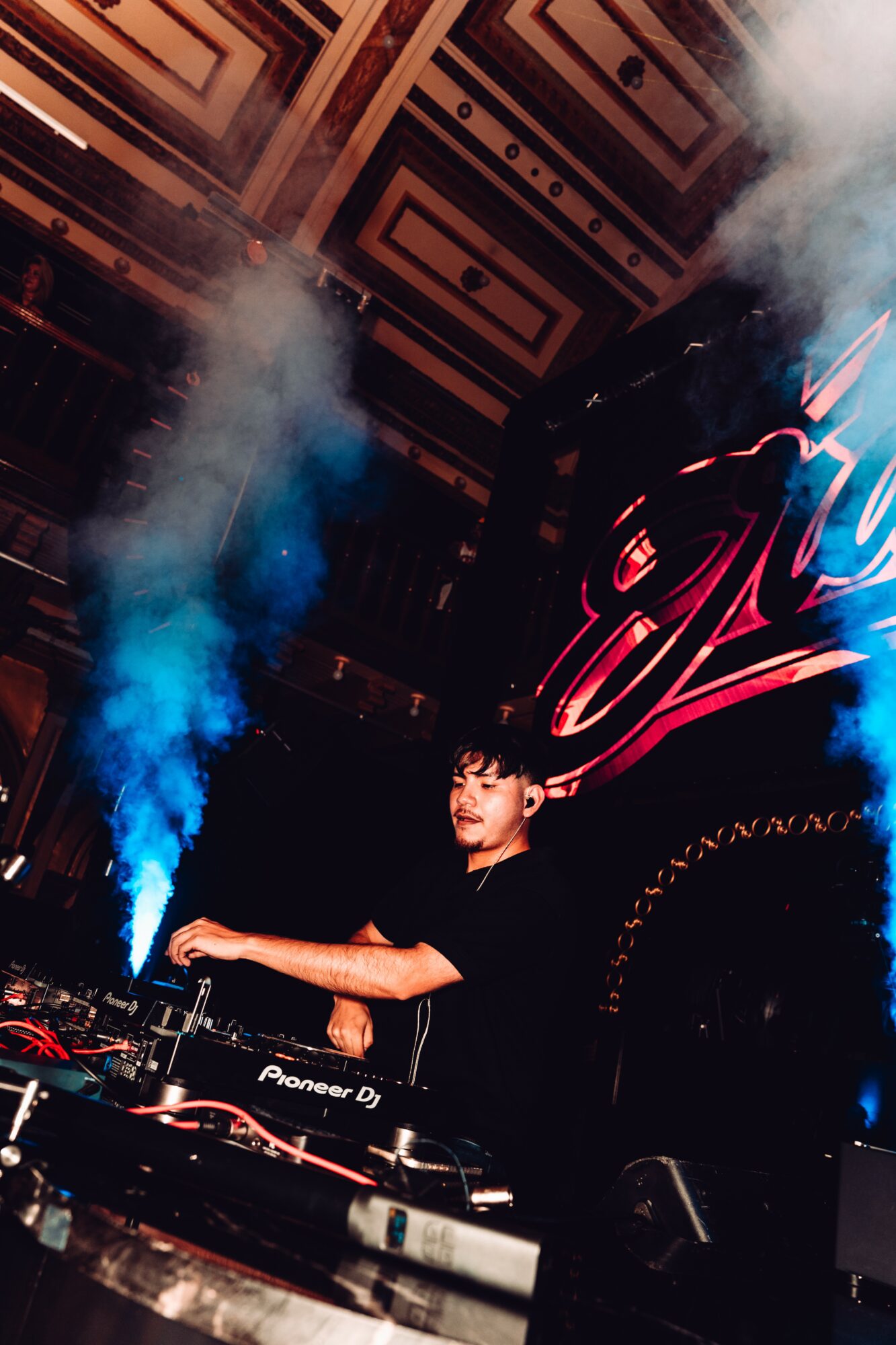 DJ performing on stage with smoke effects and colorful lighting, large decorative backdrop behind him, in a dark venue.