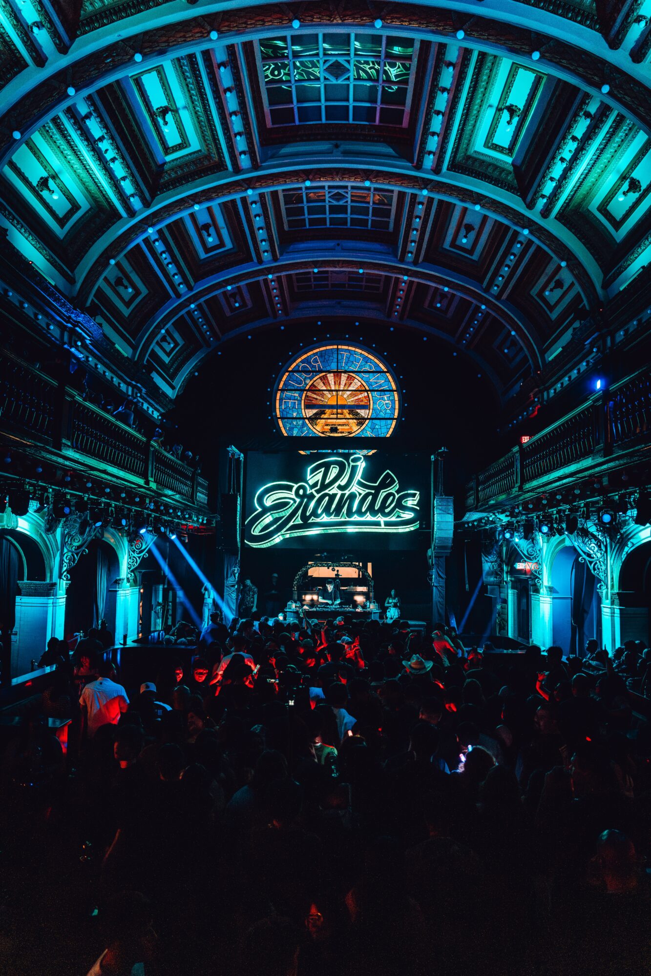 Concert stage with neon sign reading 'DJ Grandes' inside a large, ornate hall with a crowd.