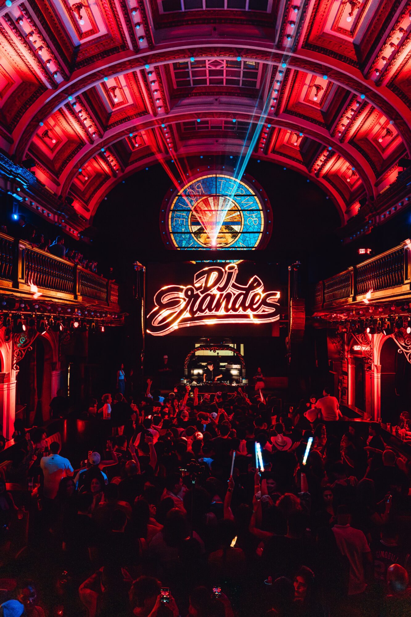 Concert venue with a high arched ceiling, red lighting, a large illuminated sign reading 'DJ Grande,' and a crowd of people.