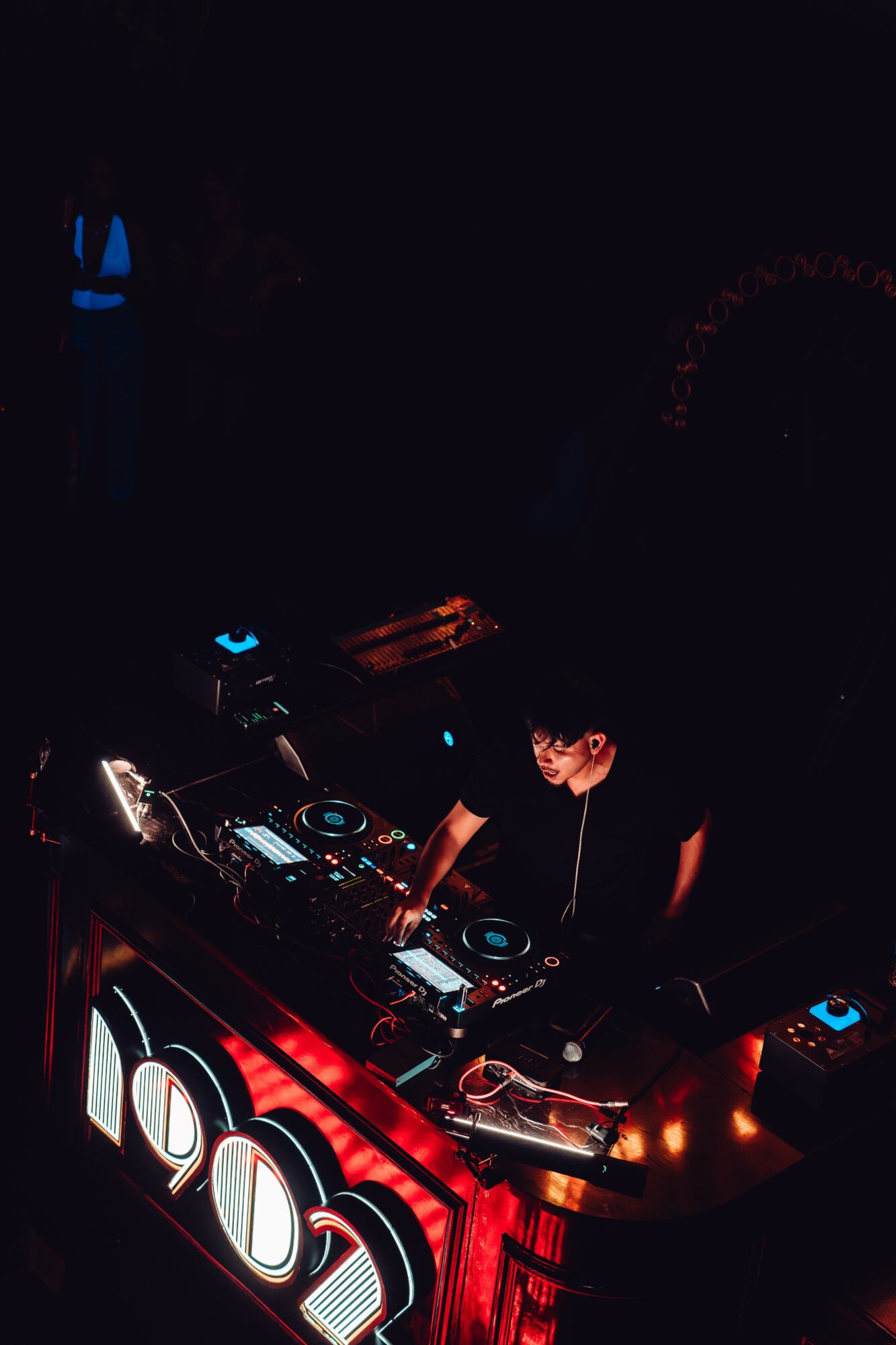 DJ performing at a dark nightclub with colorful lights and equipment, person wearing headphones, illuminated controls, and a red-lit table.