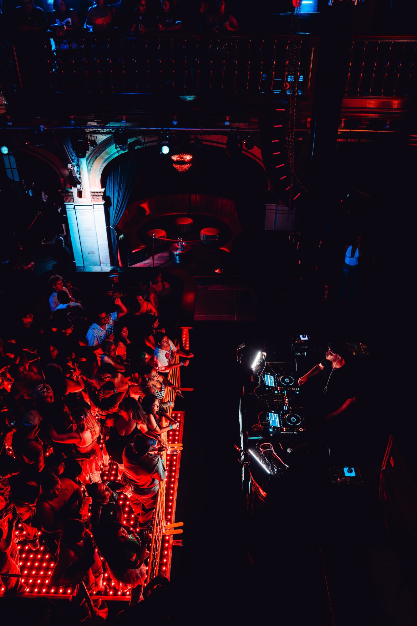 Crowd of people in a dark nightclub with red and blue lighting, DJ booth, and stage area.