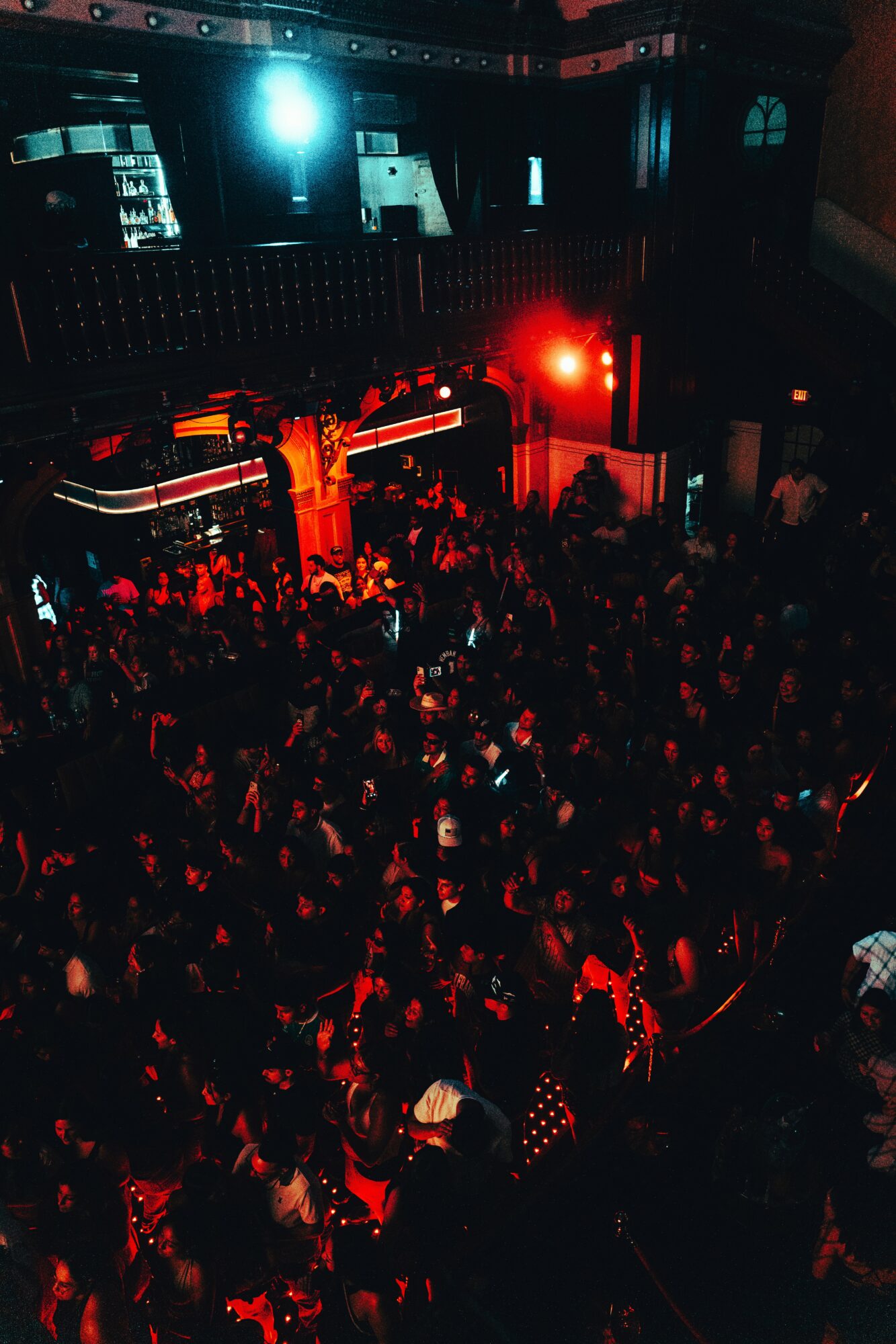 Crowd of people at a nightclub with red and blue lighting, dance floor, and bar area visible in the background.