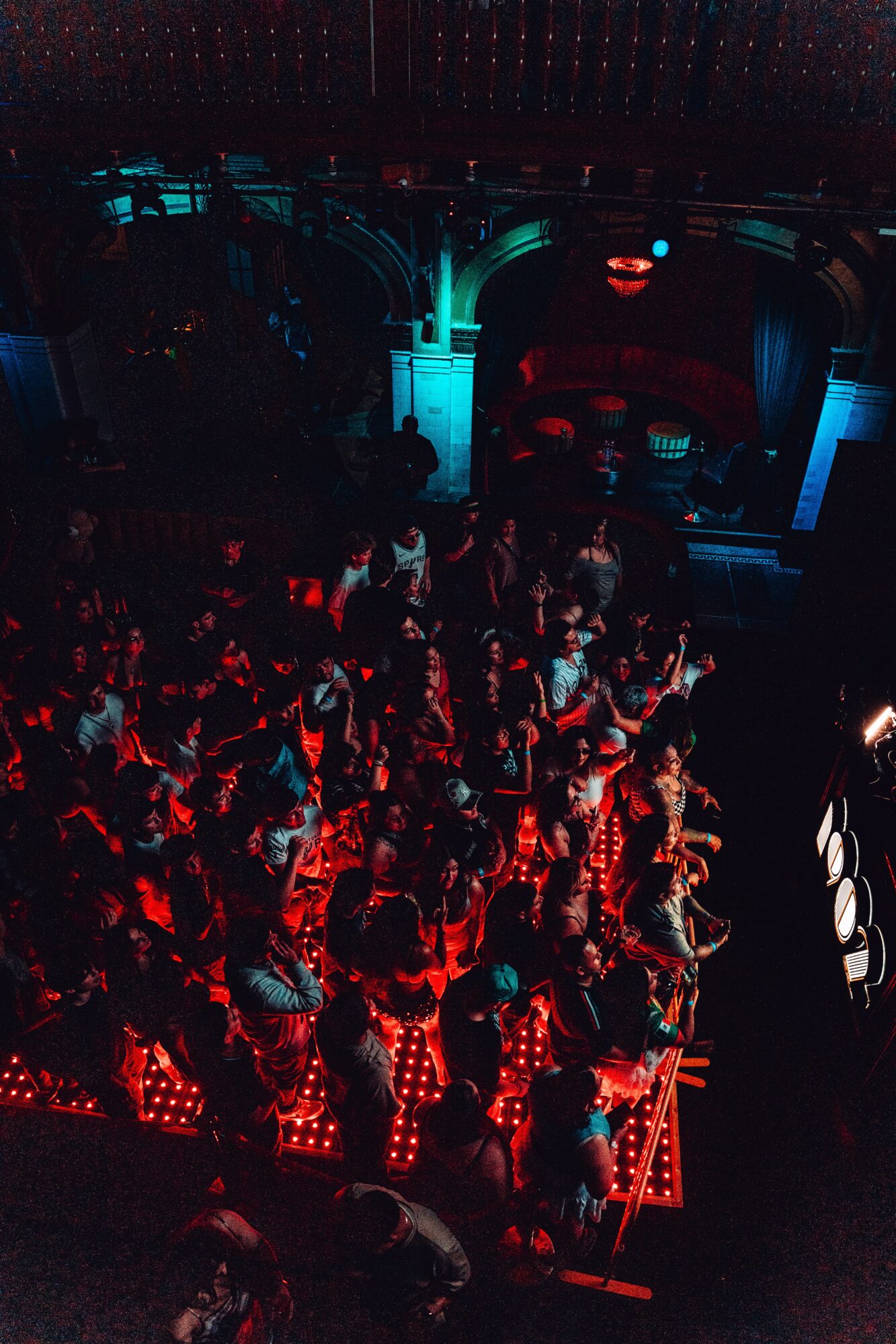 Crowd of people in a dark venue with red and blue lighting, some sitting and some standing, with a stage in the background.