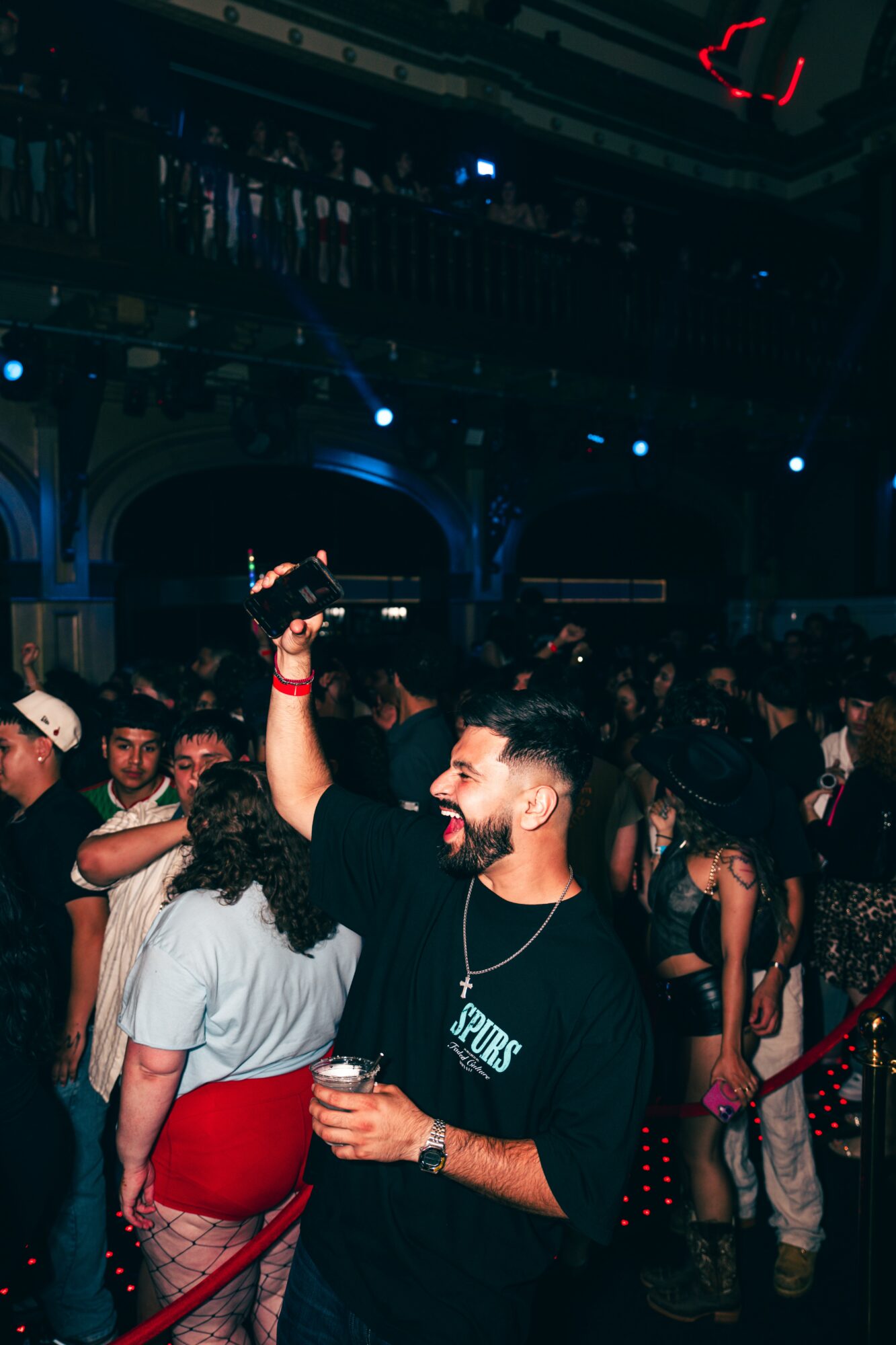 Man with a beard holding a drink and raising his hand in a crowded indoor event, smiling.