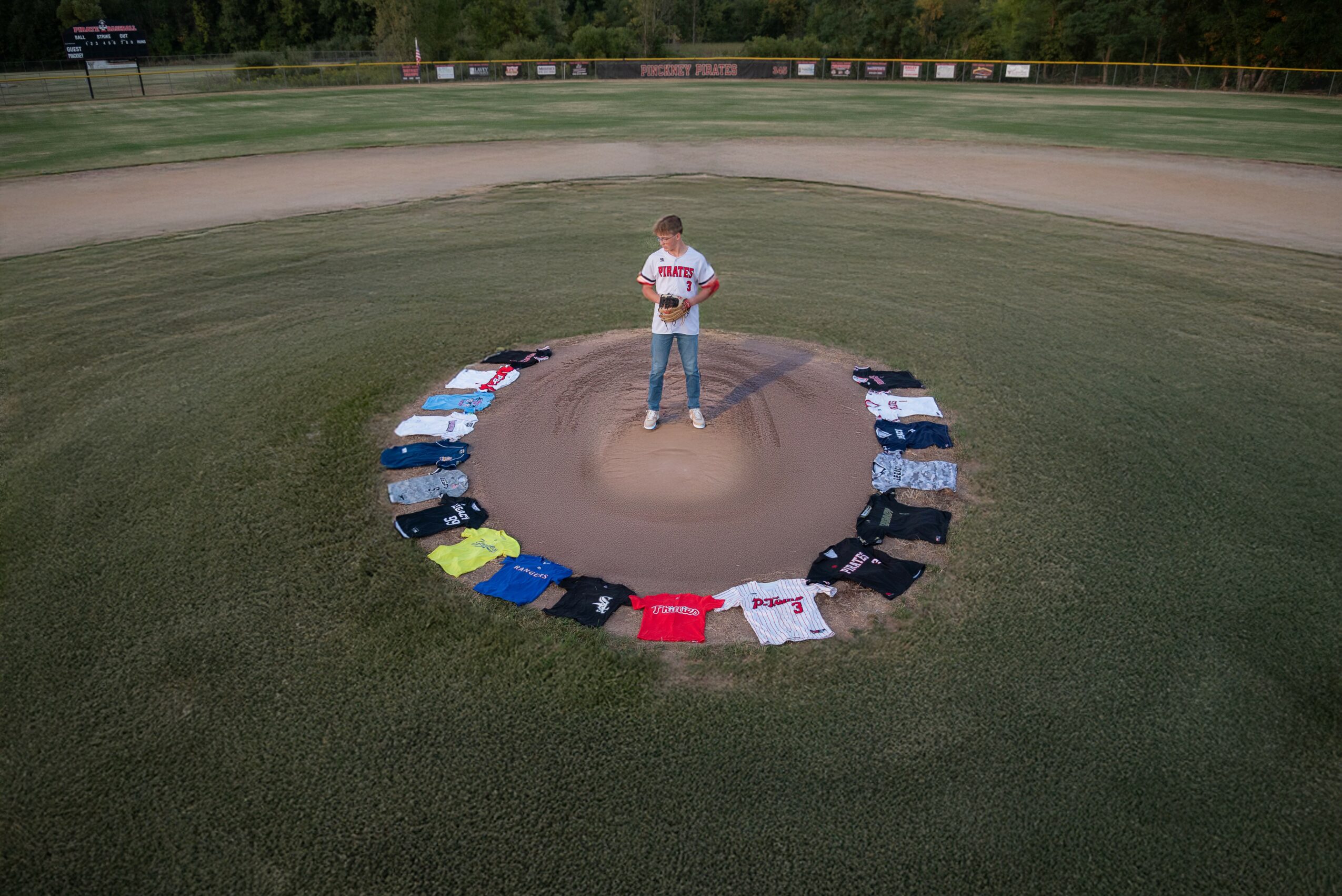 Person standing on a dirt mound in the center of a baseball field, surrounded by a circle of colorful shirts on grass.