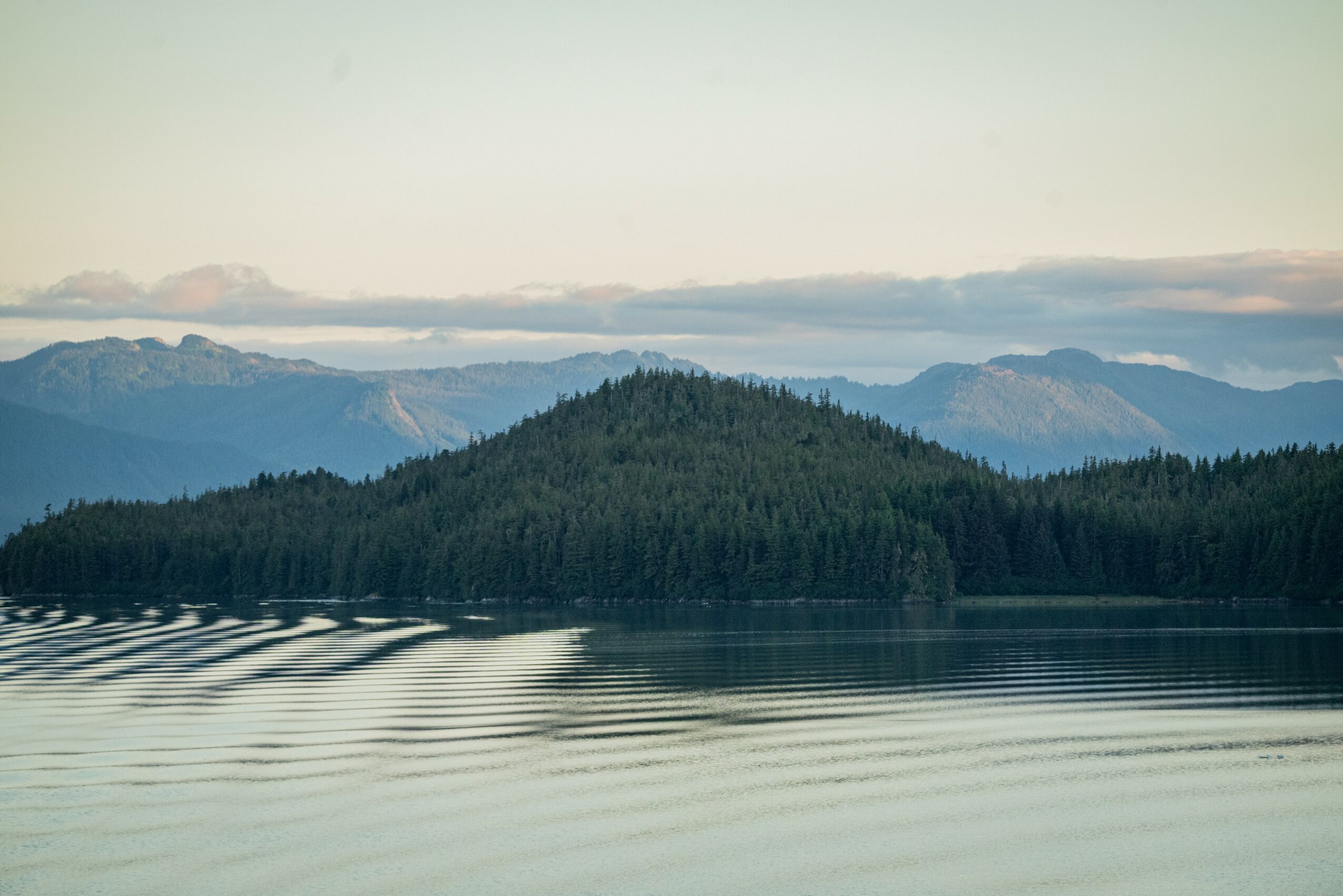 Calm lake with ripples, forested island, mountain range in background, cloudy sky, peaceful landscape