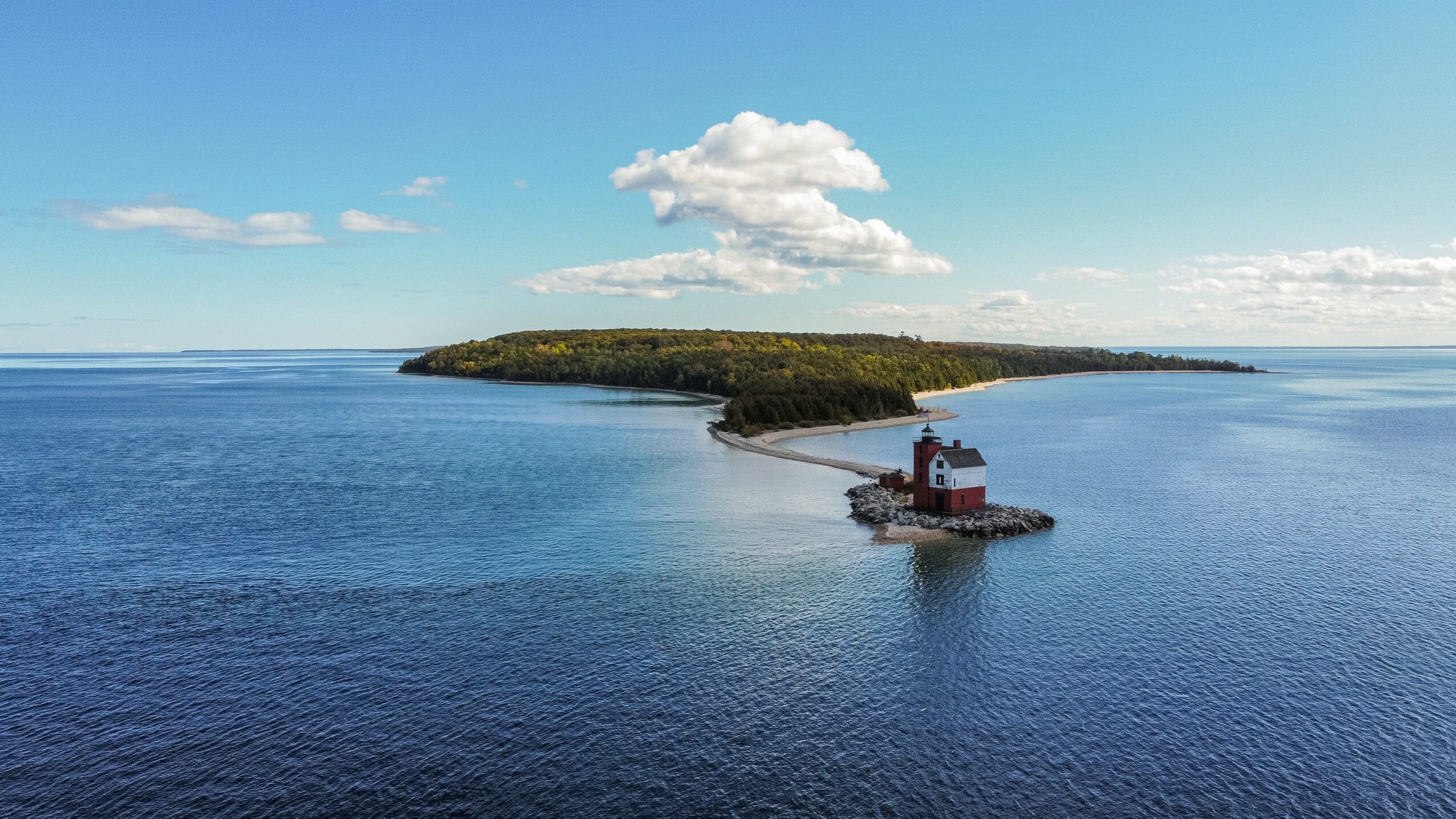 Island with a lighthouse on a small pier extending into calm blue water, under a partly cloudy sky.