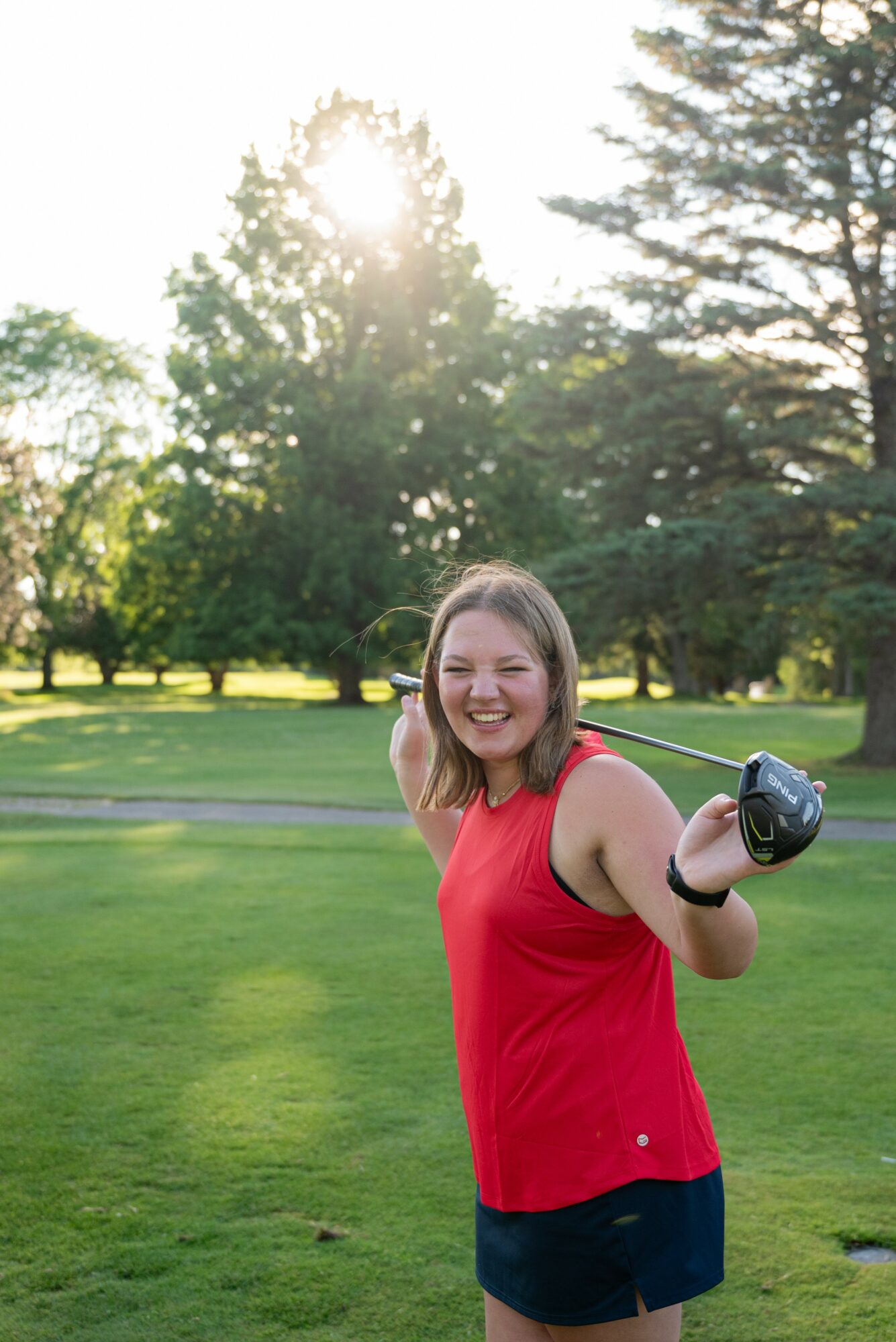 Young woman in red top and black shorts smiling outdoors holding a golf club, trees and sunlight in background.