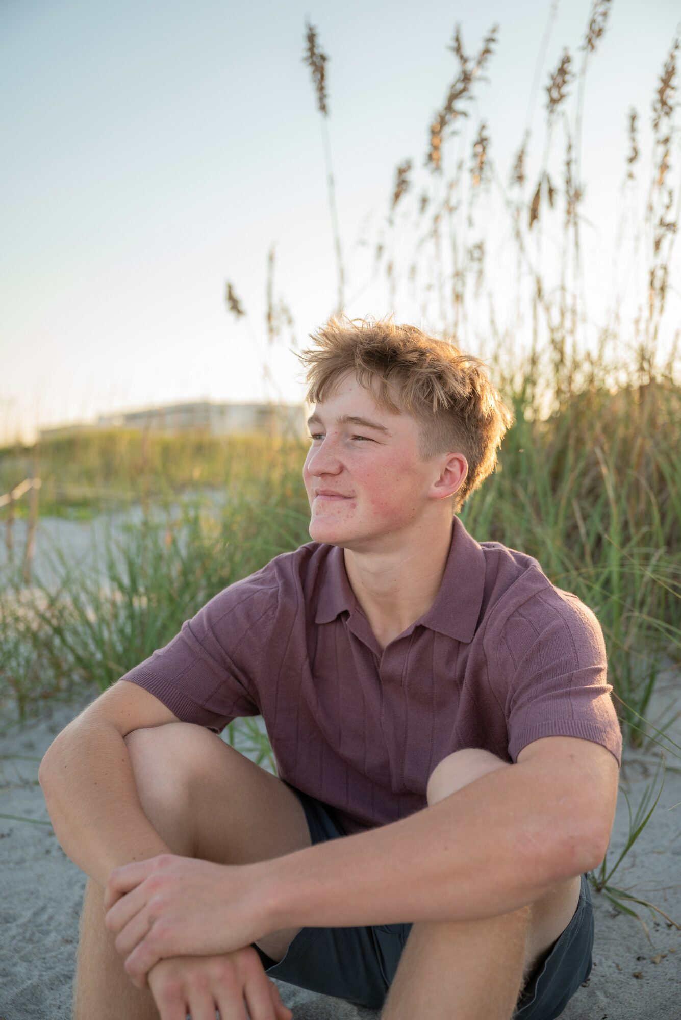 Young man sitting outdoors near tall grass, smiling and looking to the side, during sunset.