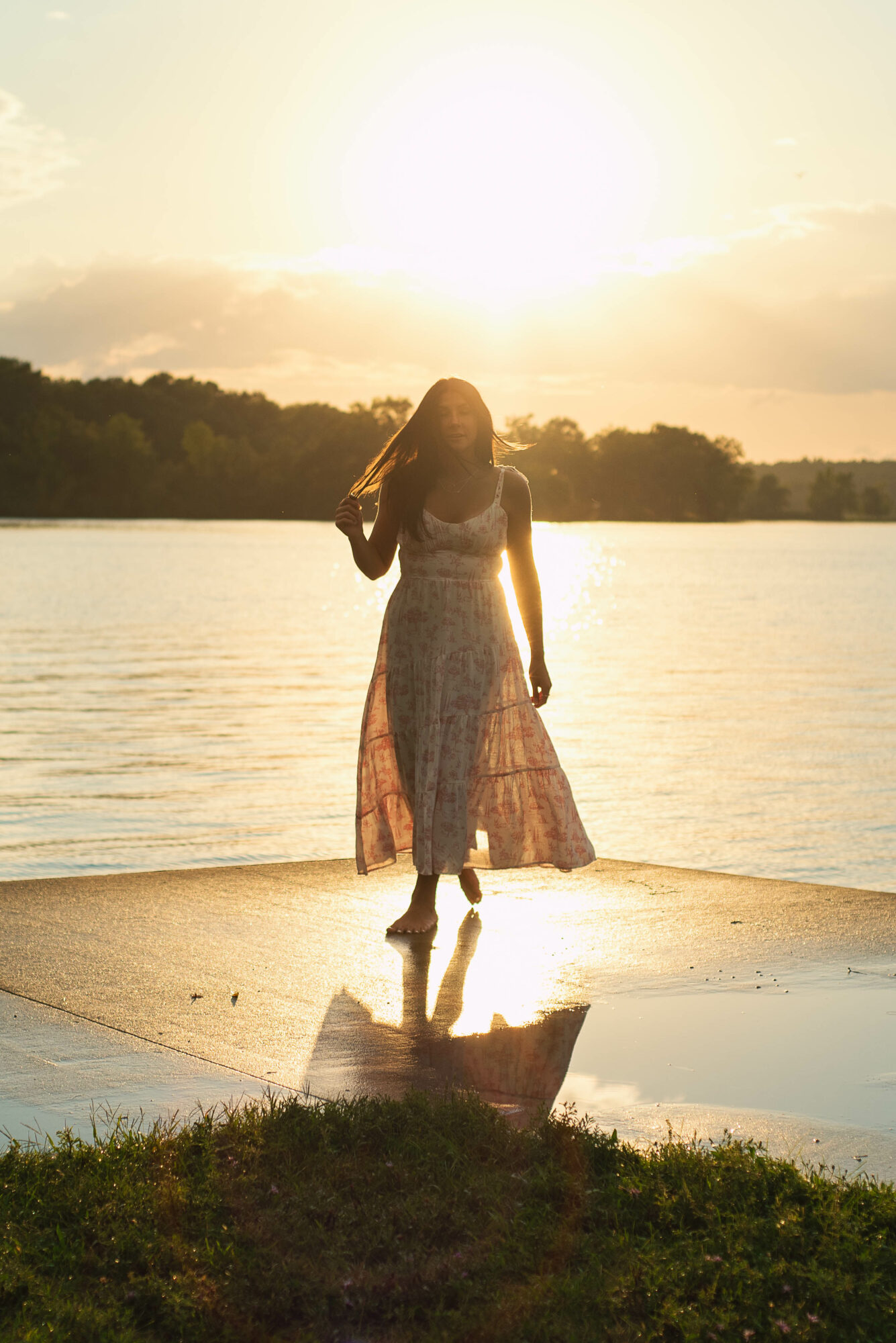 Person in a long dress standing on a dock by water during sunset, with reflection visible in the water.