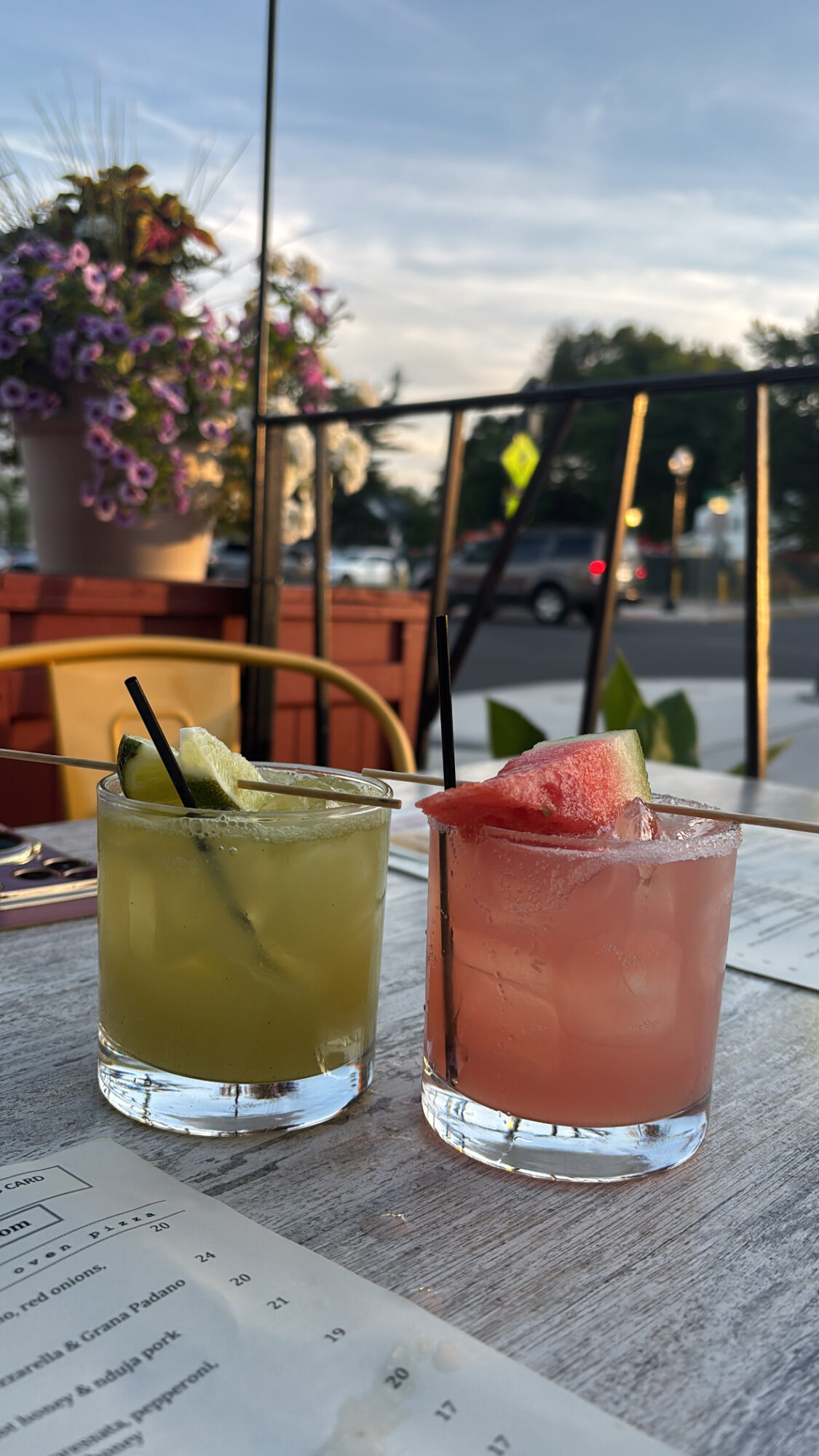 Two colorful cocktails with ice and straws on a table outdoors, with flowers and trees in the background.