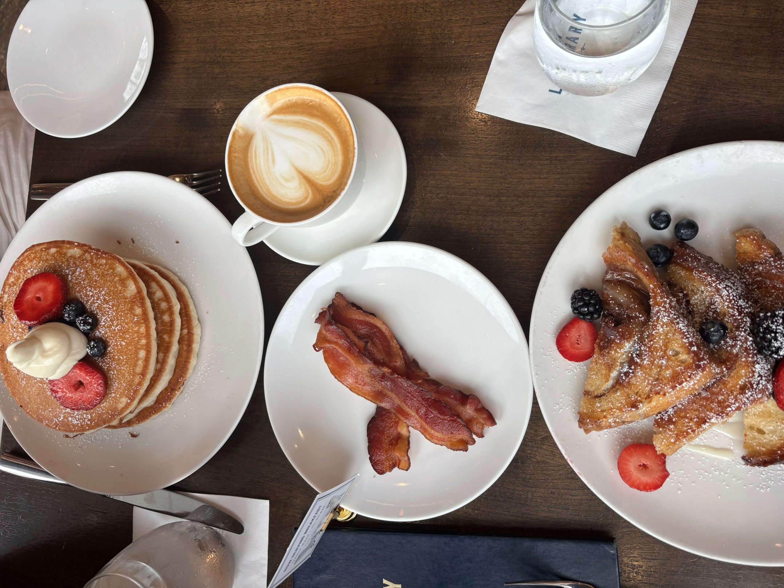 Pancakes with berries and whipped cream, bacon, and a waffle with fruit on plates, coffee, and water on a wooden table.