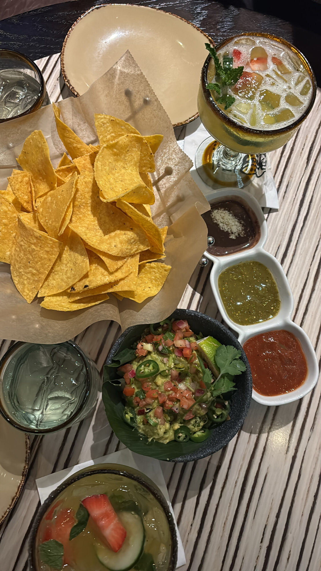Bowl of tortilla chips, margarita, and various dips on a table, with a glass of water and a bowl of guacamole.