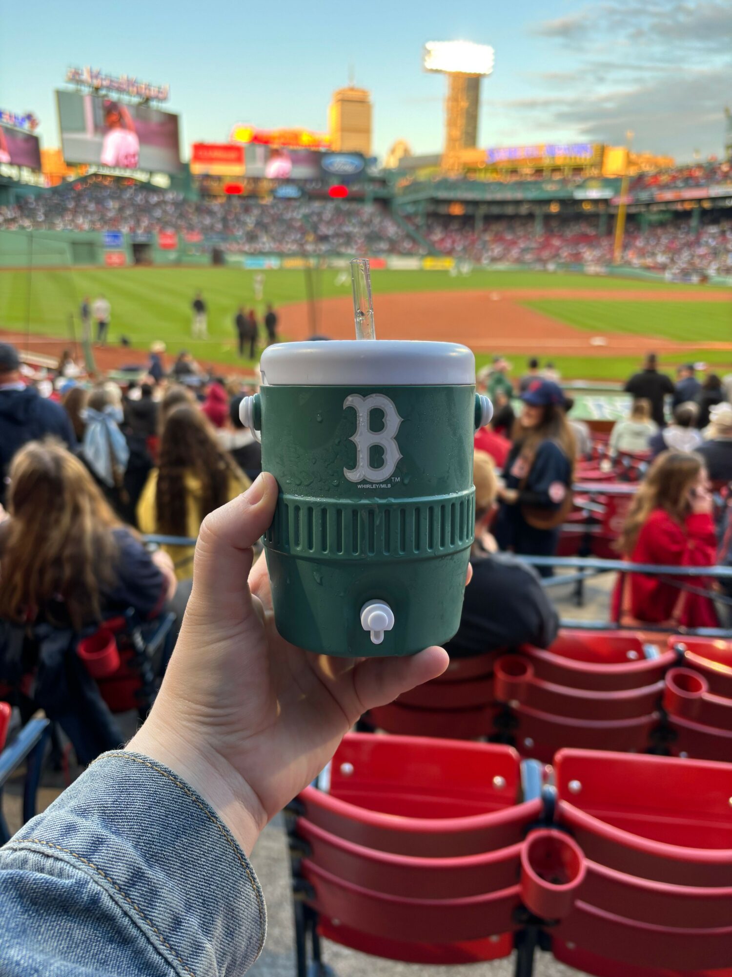 Person holding a green and white B beer cup at a baseball game stadium with red seats and spectators.