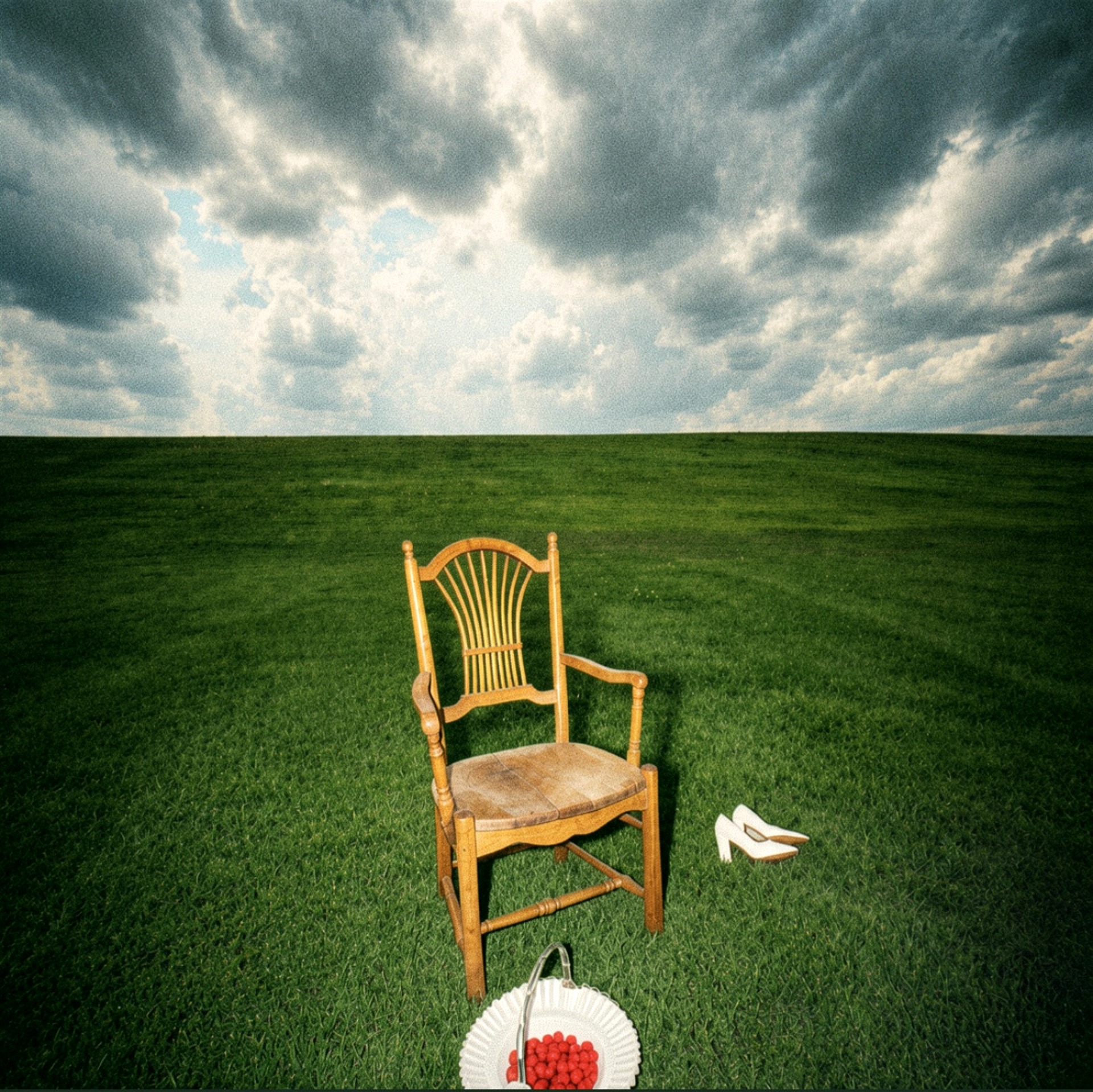 A wooden chair and a pair of white shoes on a grassy field under a cloudy sky.