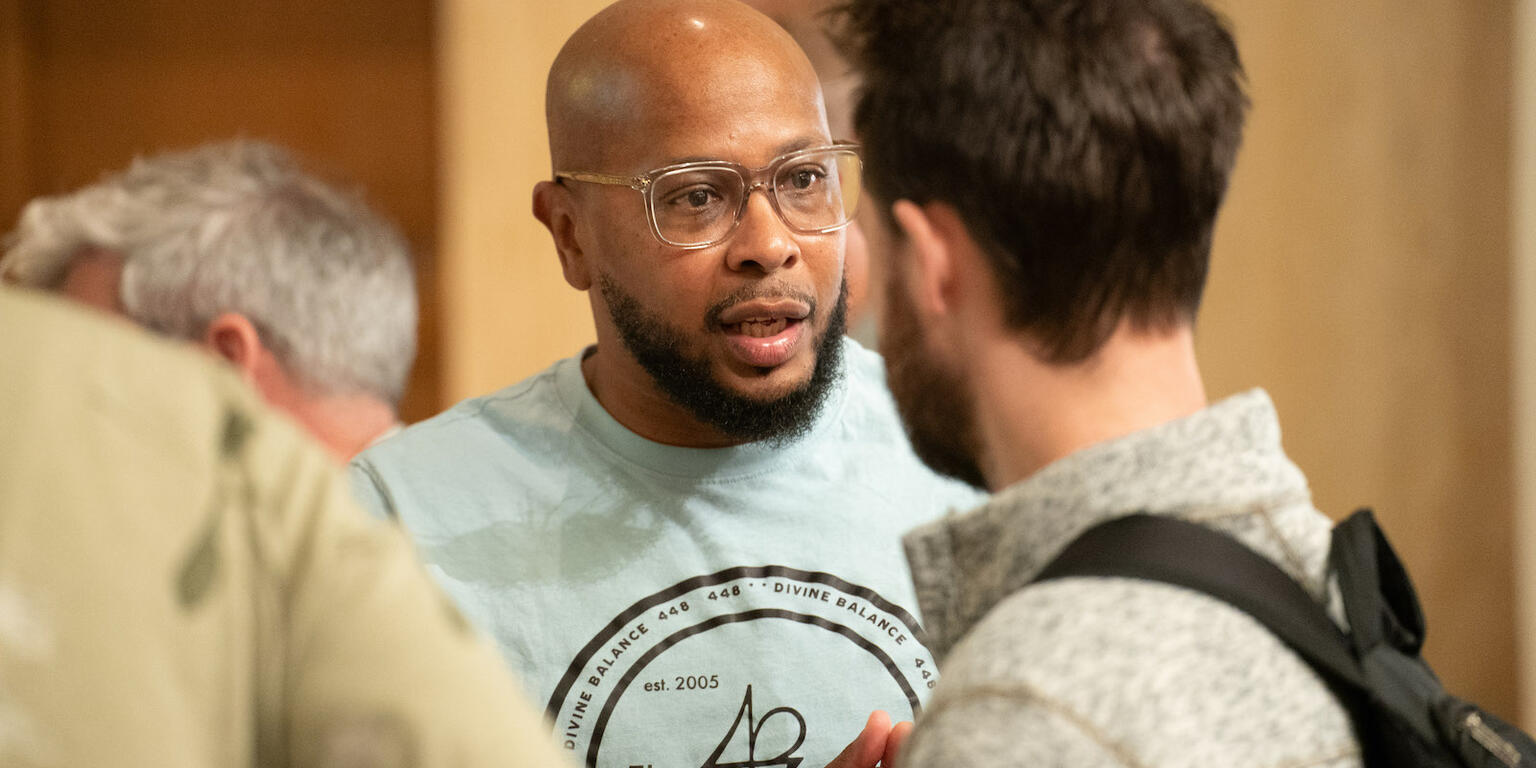 Two men engaged in conversation indoors, one with glasses and a beard, the other with dark hair and a backpack.